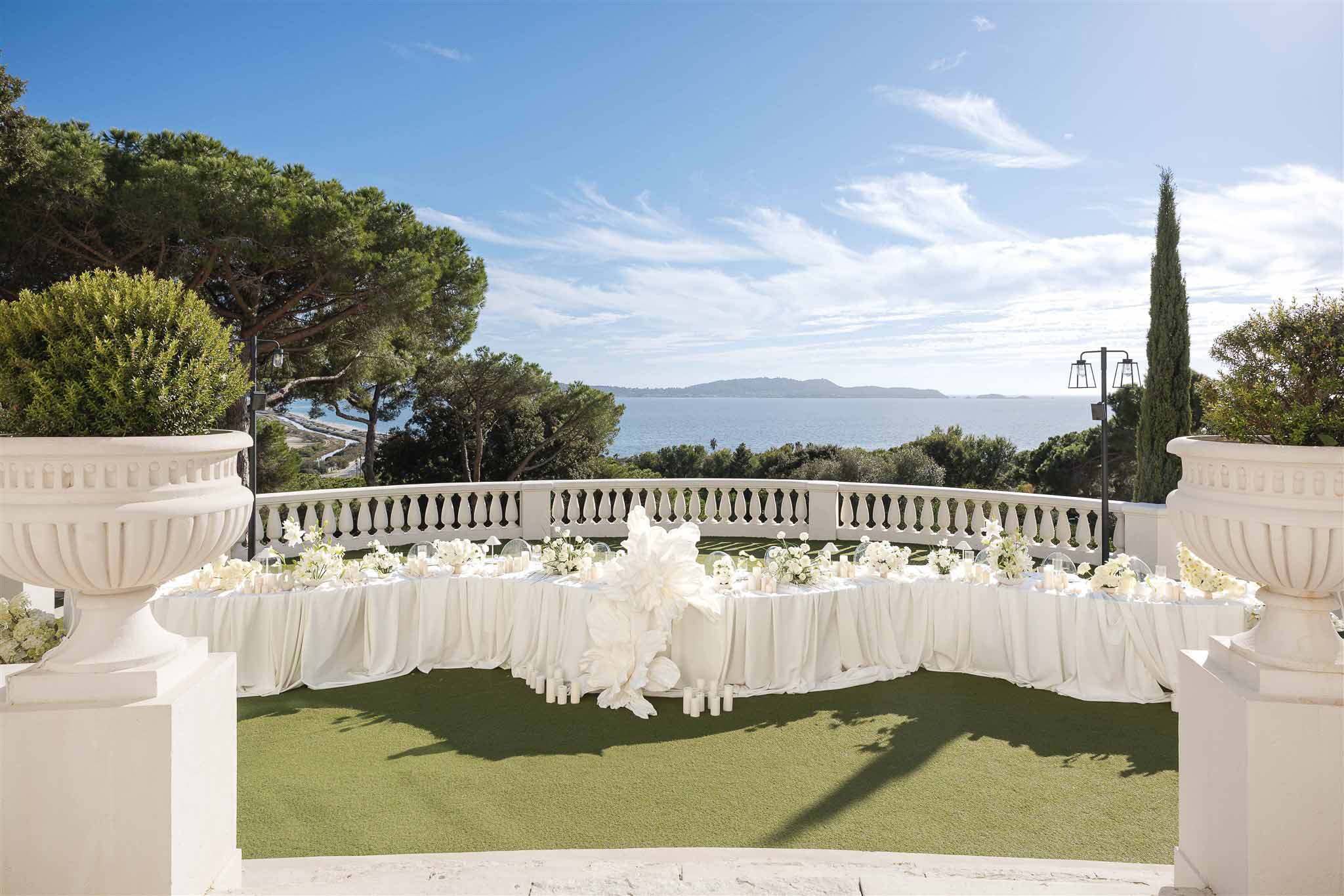 All-white head table with orchid and rose clusters and pillar candles on sea-view terrace with urns