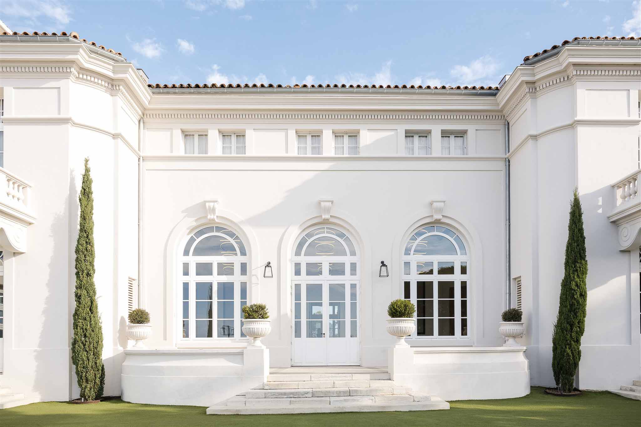 White rendered villa with three arched doors, columnar cypress trees, and topiary urn planters on steps