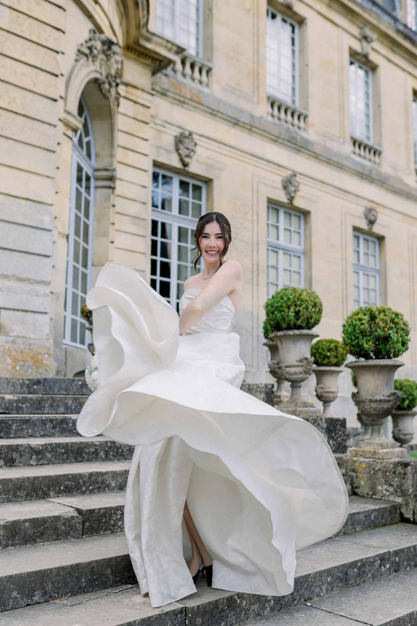 Bride twirling strapless ballgown on chateau stone steps with topiary urns and arched windows behind
