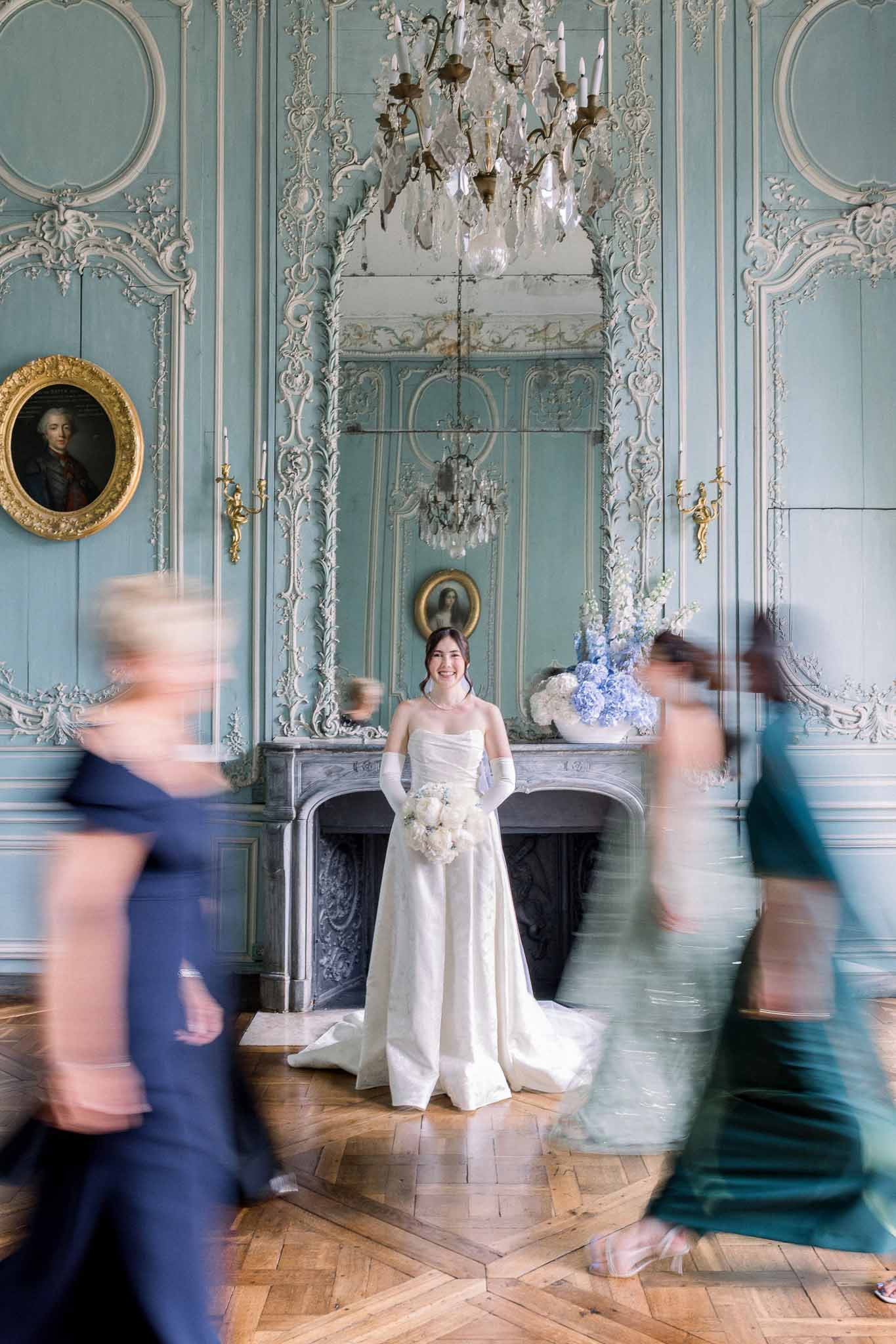 Bride in strapless ballgown with white gloves in Rococo salon as two guests pass in motion blur