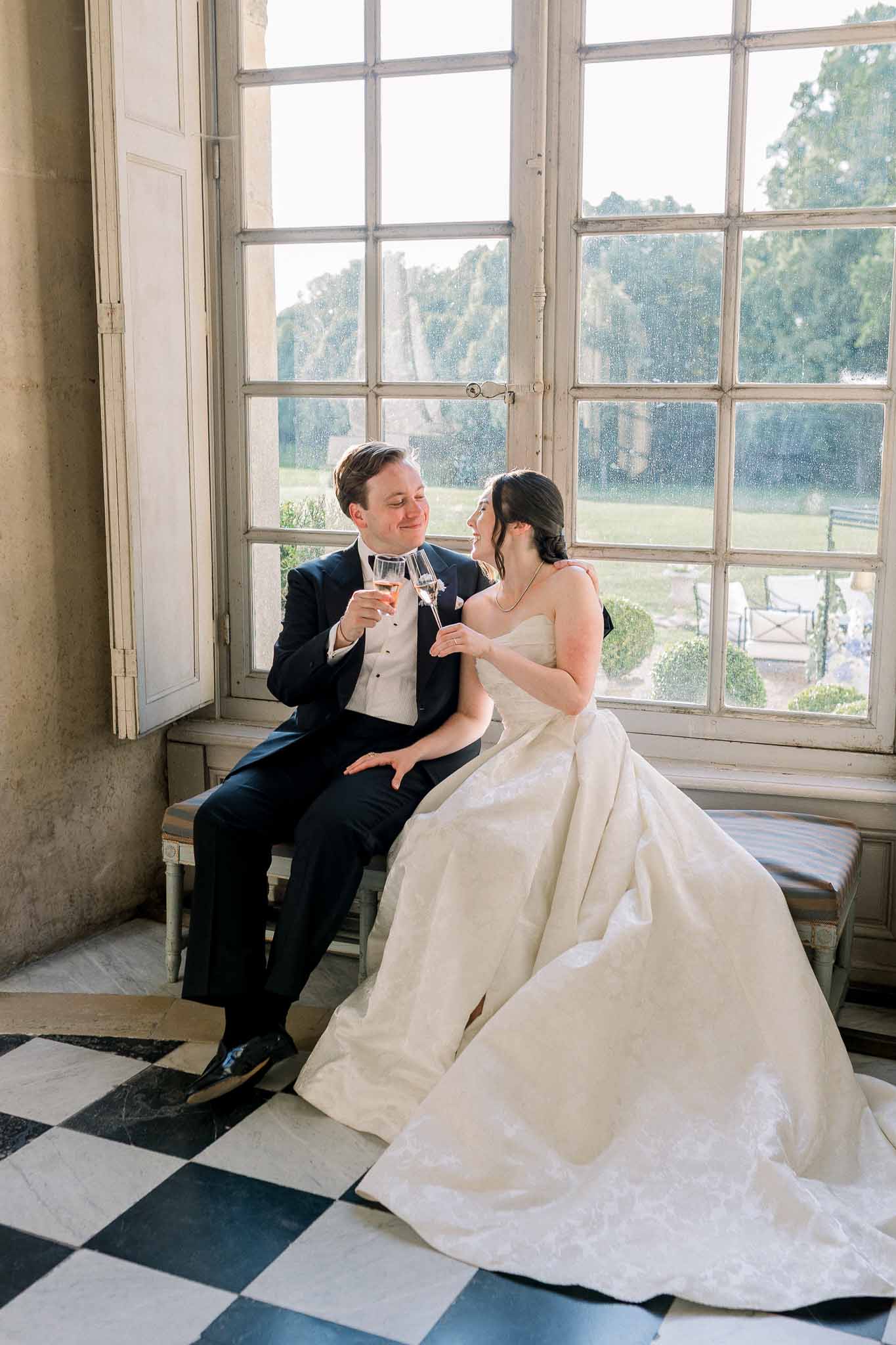 Bride in ivory ballgown and groom in navy tuxedo clinking champagne by multi-pane window overlooking gardens