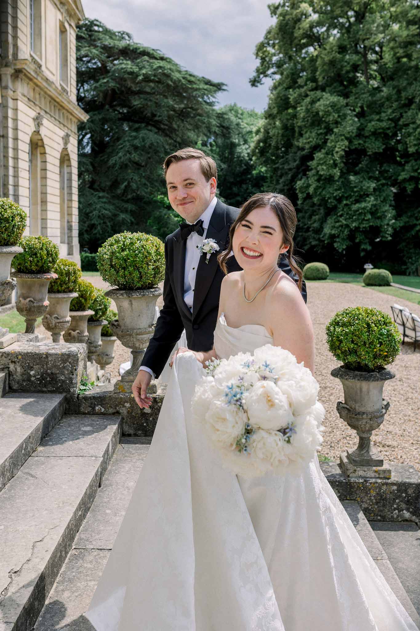 Laughing bride and groom on stone chateau terrace steps with white peony bouquet and formal topiary gardens