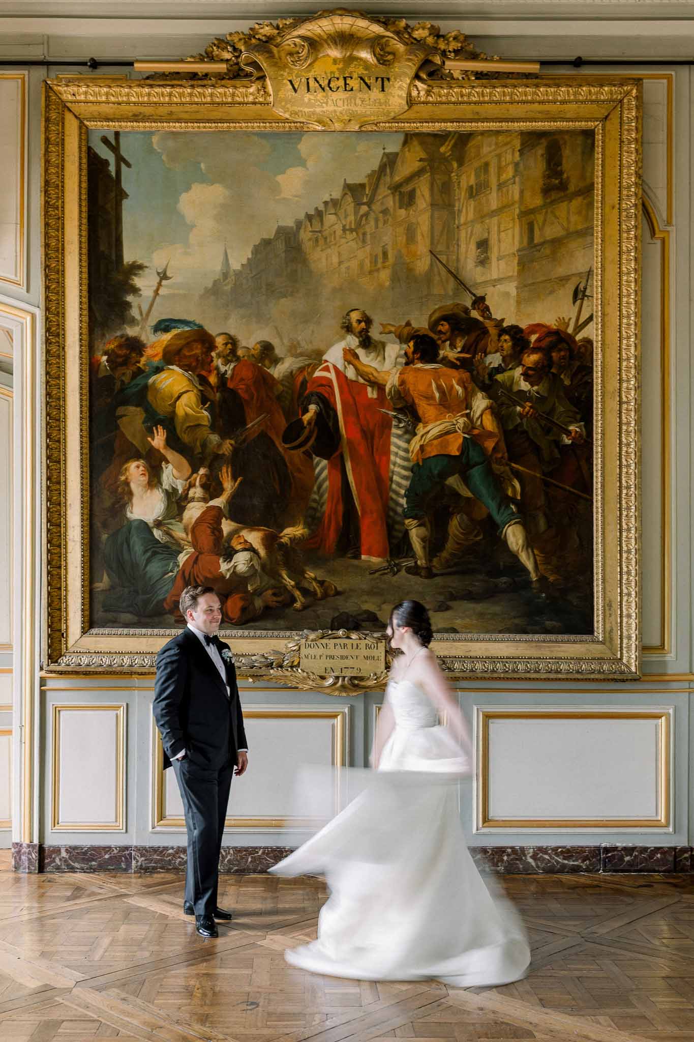 Bride in white strapless ballgown and groom in black tuxedo posing before a gilt-framed painting inside a grand Parisian i...