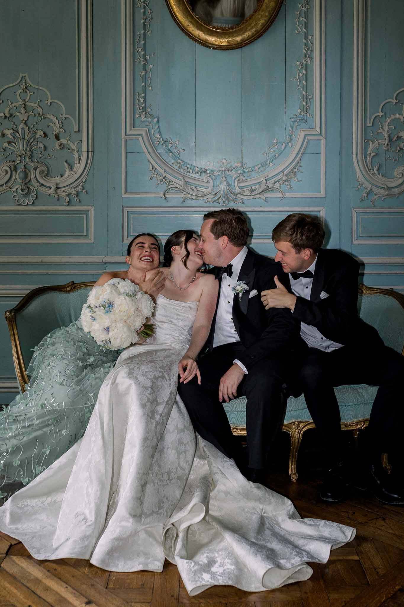 Bride groom and guests laughing on a gold settee in a powder blue paneled French chateau salon