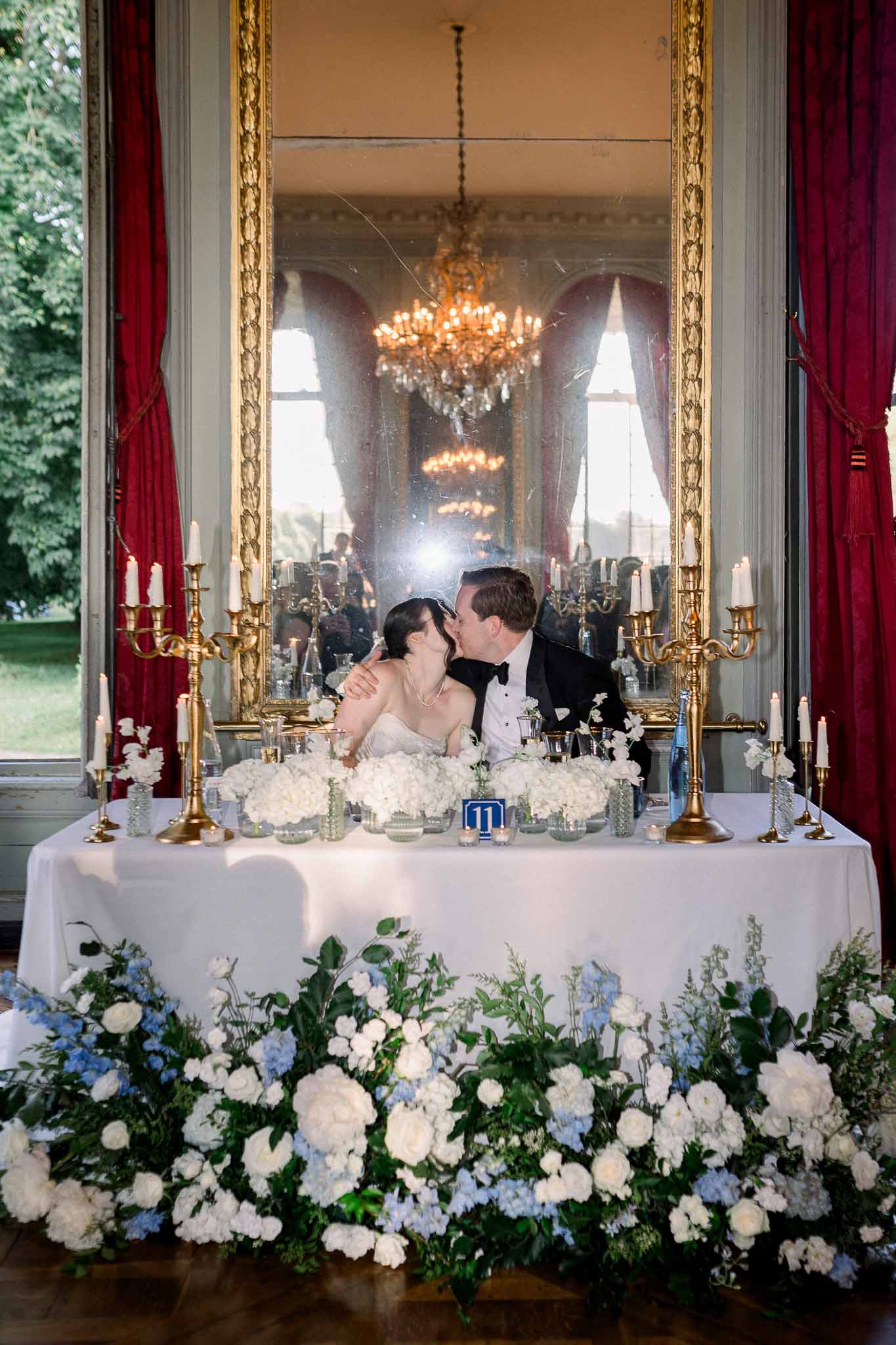 Bride and groom kiss at sweetheart table with white and blue hydrangeas in crimson-curtained ballroom