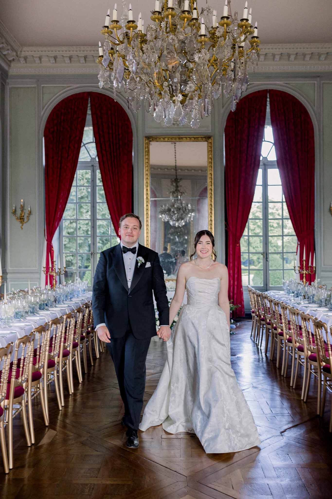 Bride in jacquard ballgown and groom in tuxedo walk between reception tables in sage-panelled ballroom