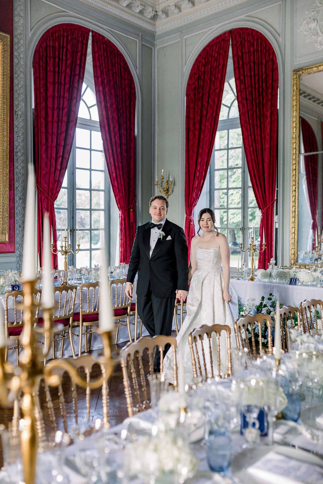 Bride and groom walking between banquet tables in panelled ballroom with crimson drapes and gold chairs
