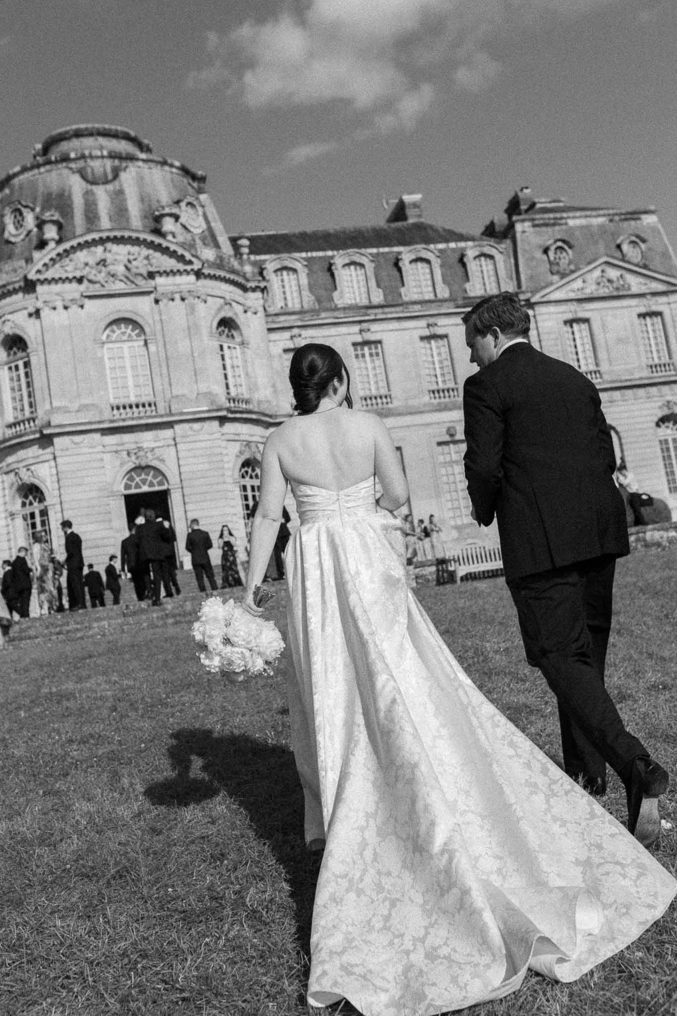 Black and white rear view of bride and groom walking across lawn toward Baroque chateau with guests on terrace
