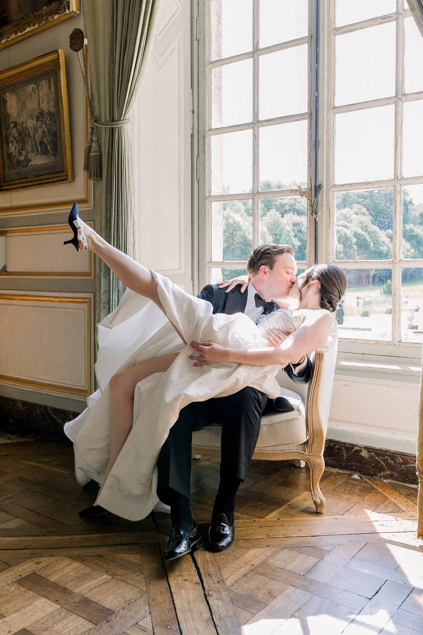 Bride sits on groom's lap in Louis XVI chair kissing in panelled chateau salon with parquet floor