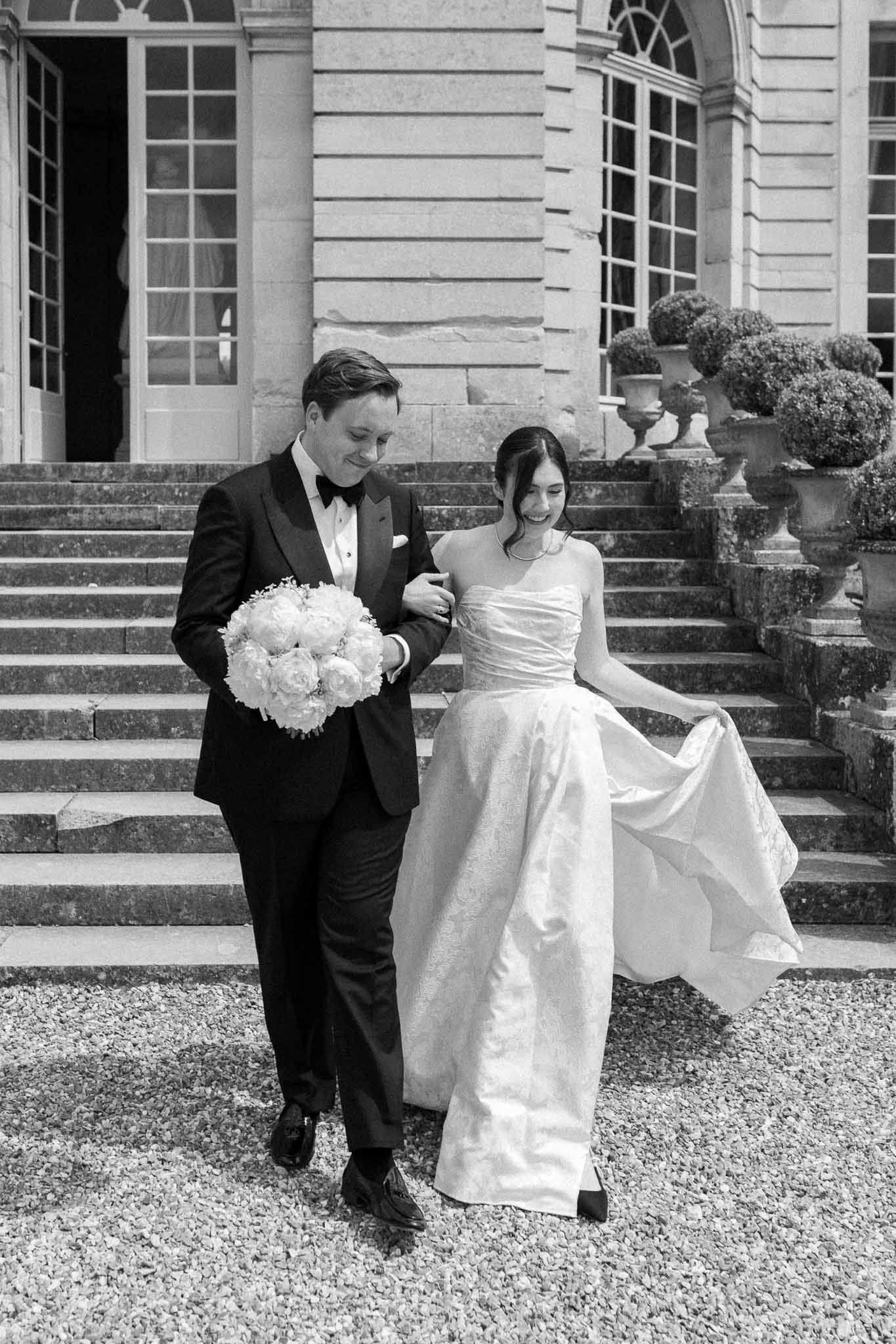 Black and white couple on gravel at chateau staircase, bride laughing in ballgown, groom carrying bouquet