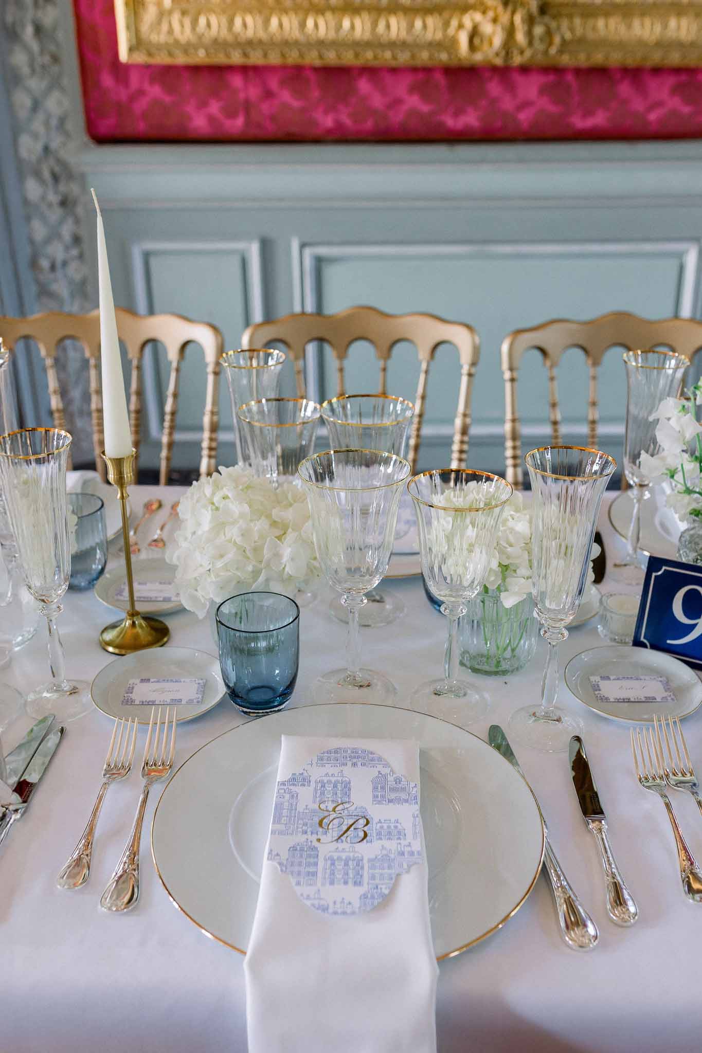 Blue-and-gold table setting with toile menu cards, white hydrangea, navy glassware, and gold Napoleon chairs