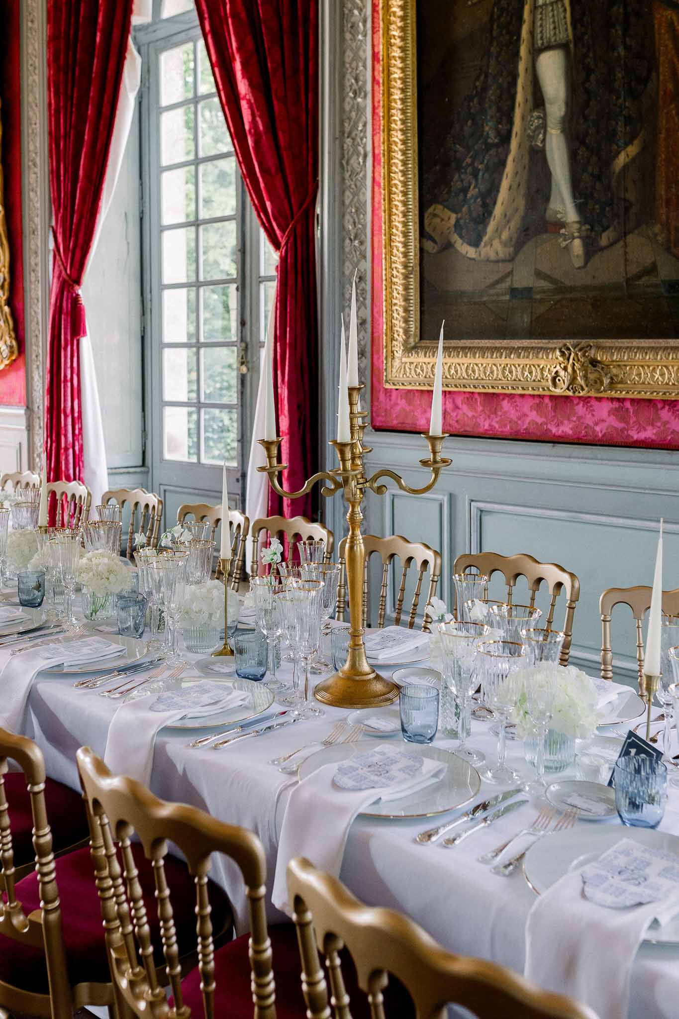 Formal chateau reception table with brass candelabra, white hydrangeas, and blue glassware by crimson drapes