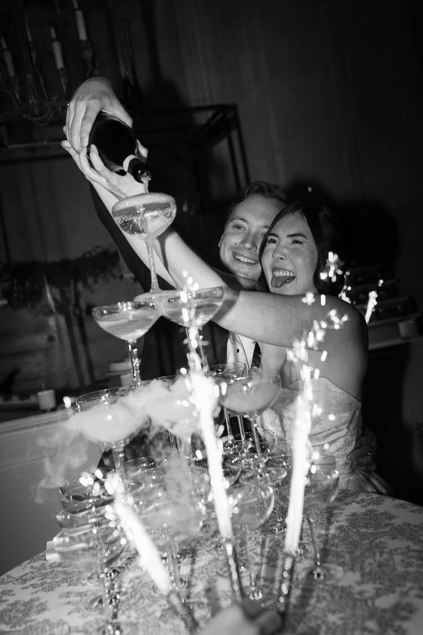 Bride and groom pour champagne tower with sparklers and dry ice smoke at indoor reception in B&W