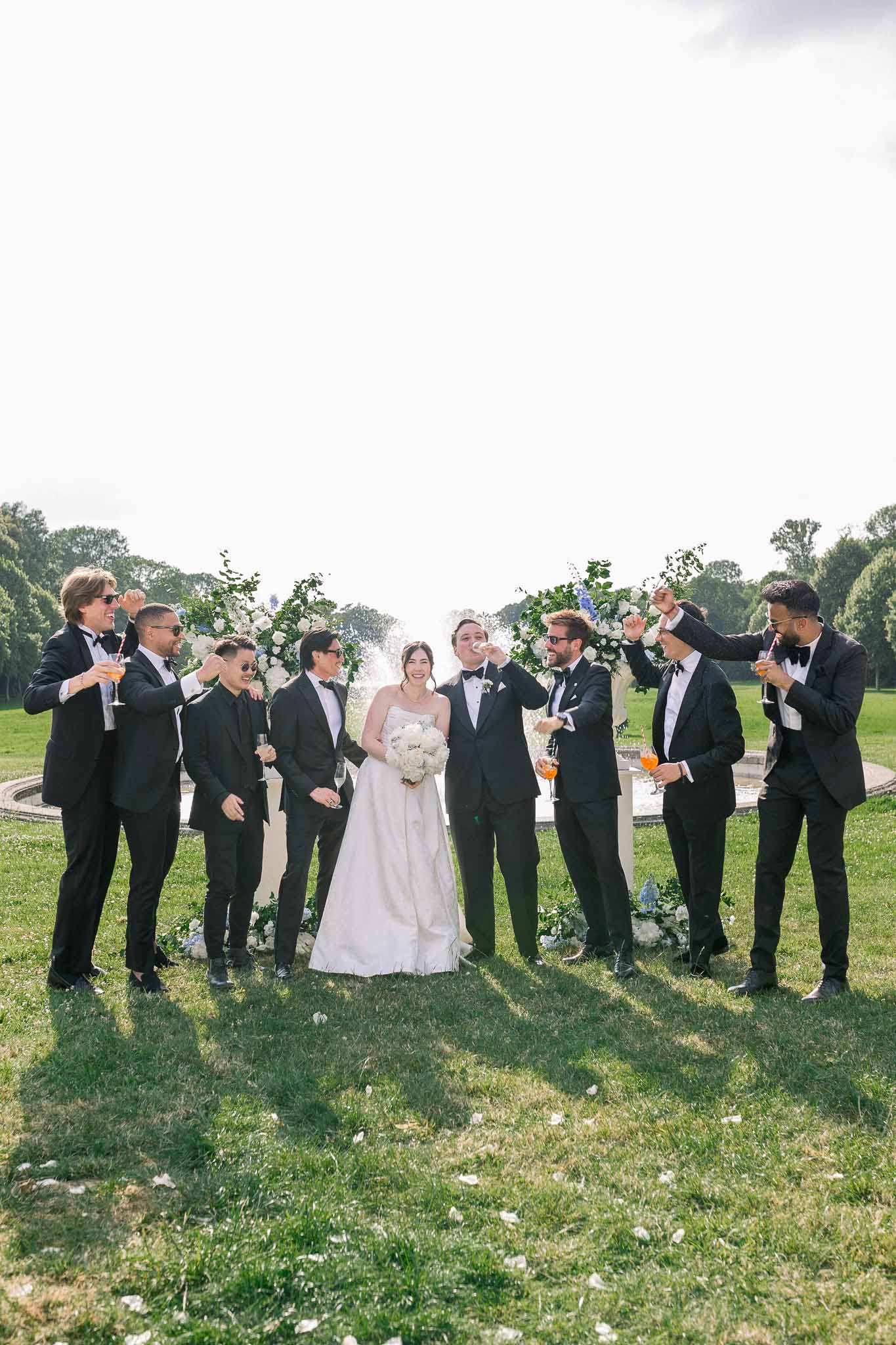 Bride and seven groomsmen in black tuxedos raising drinks before white and blue floral ceremony arch