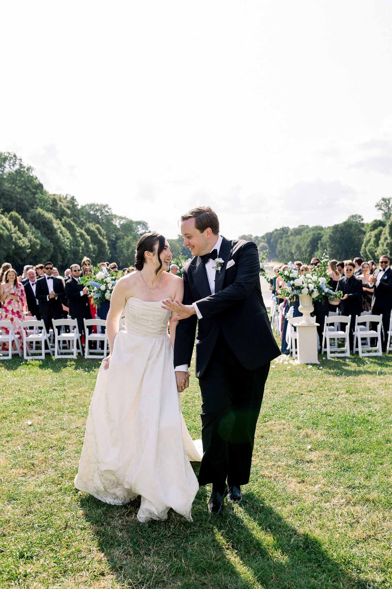 Bride and groom walk back down outdoor garden aisle as guests applaud, white rose and blue hydrangea arrangements