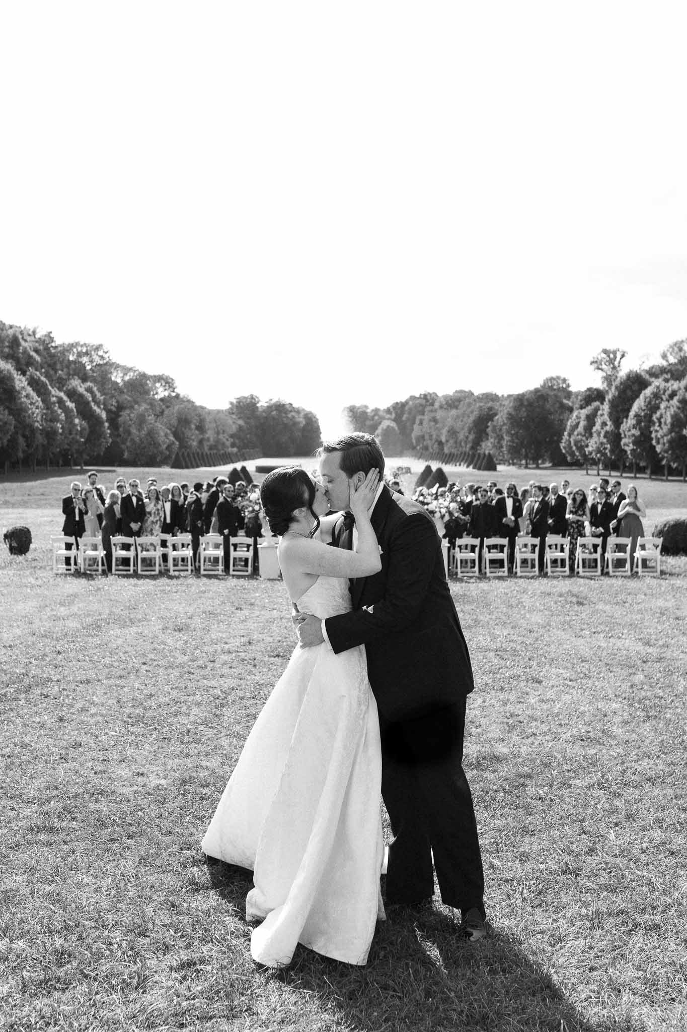 First kiss on formal tree-lined allee with 70 seated guests in white folding chairs in B&W