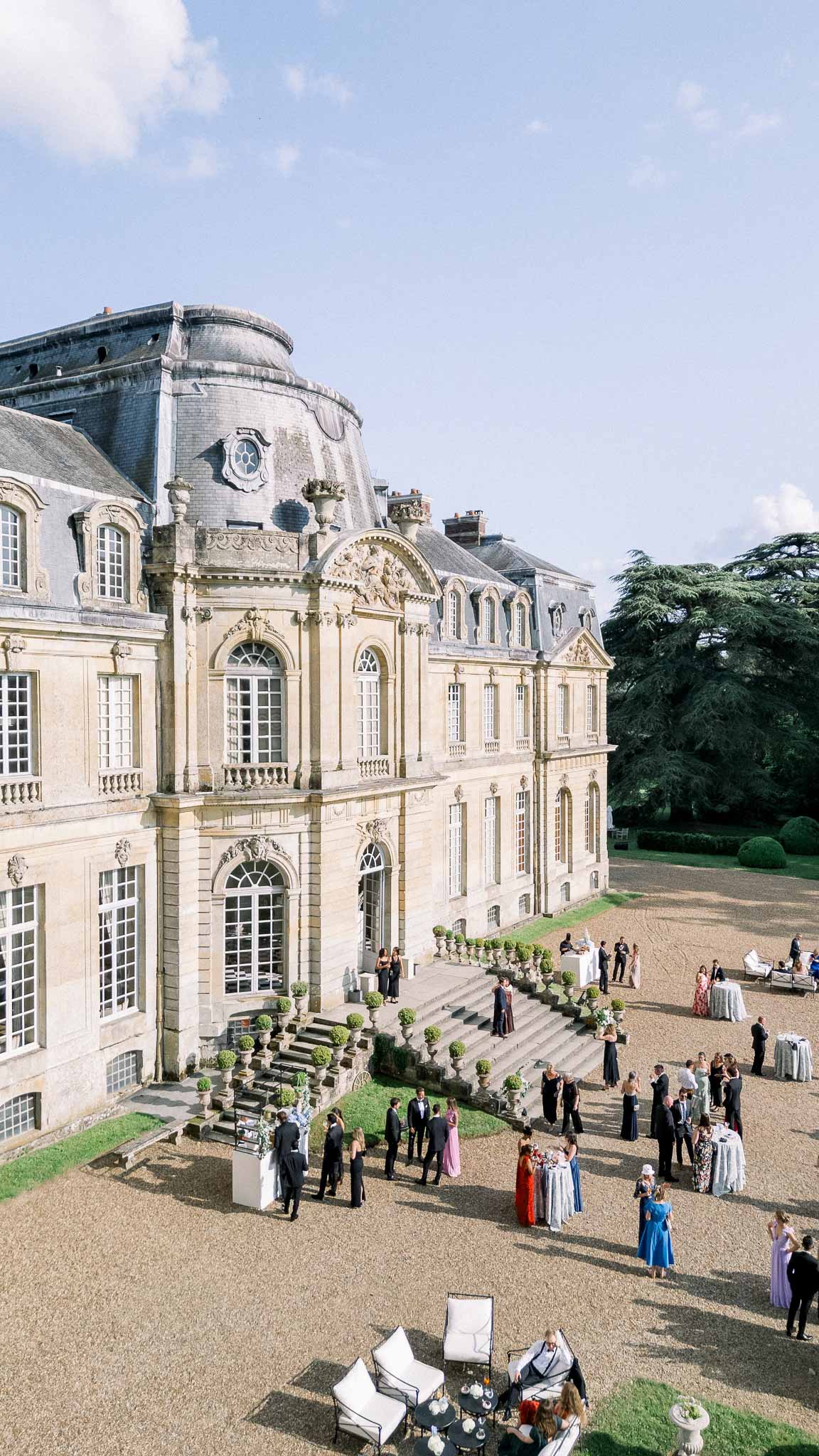 Aerial view of cocktail hour on gravel terrace of a grand French chateau with guests and white lounge furniture