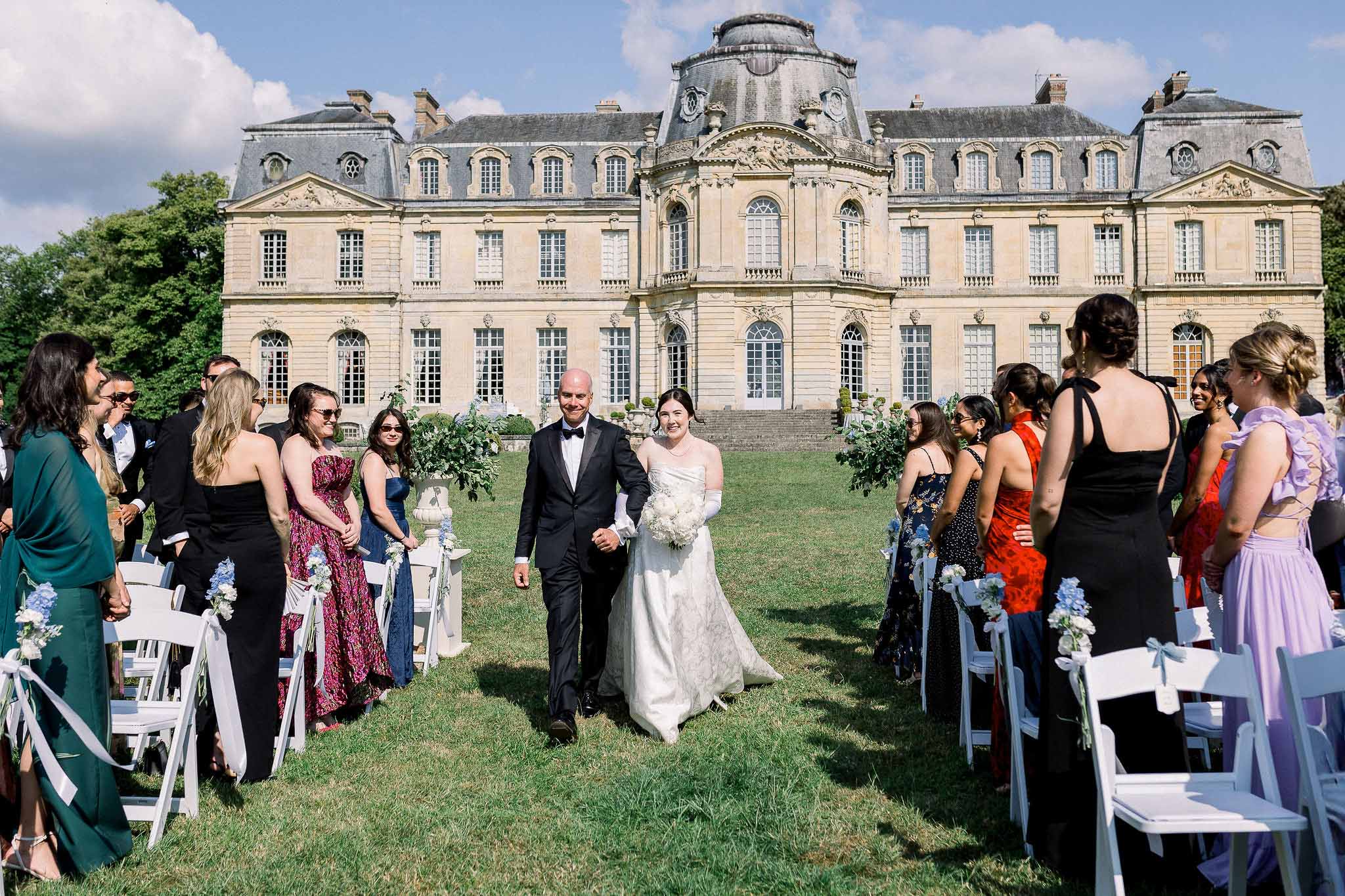 Bride and groom recess down a lawn aisle lined with white chairs and blue delphinium at a chateau