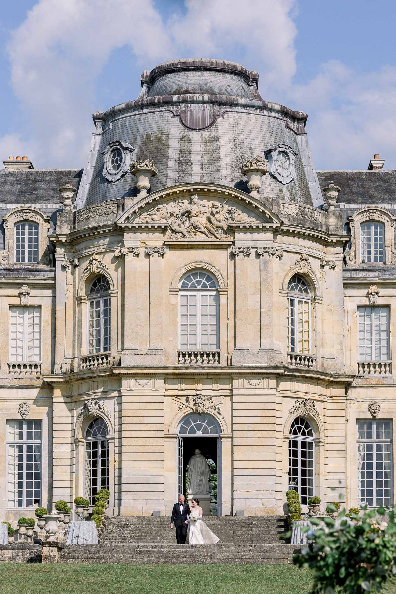Couple descending grand stone staircase of baroque French chateau, bride in ivory ballgown and groom in black tuxedo