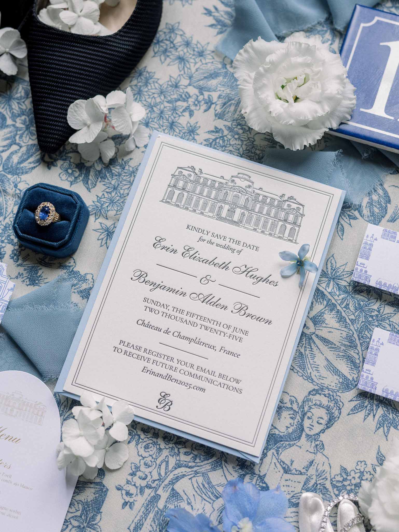 Flat lay of blue and white wedding stationery suite with save-the-date card, sapphire ring, and hydrangea blooms on toile ...