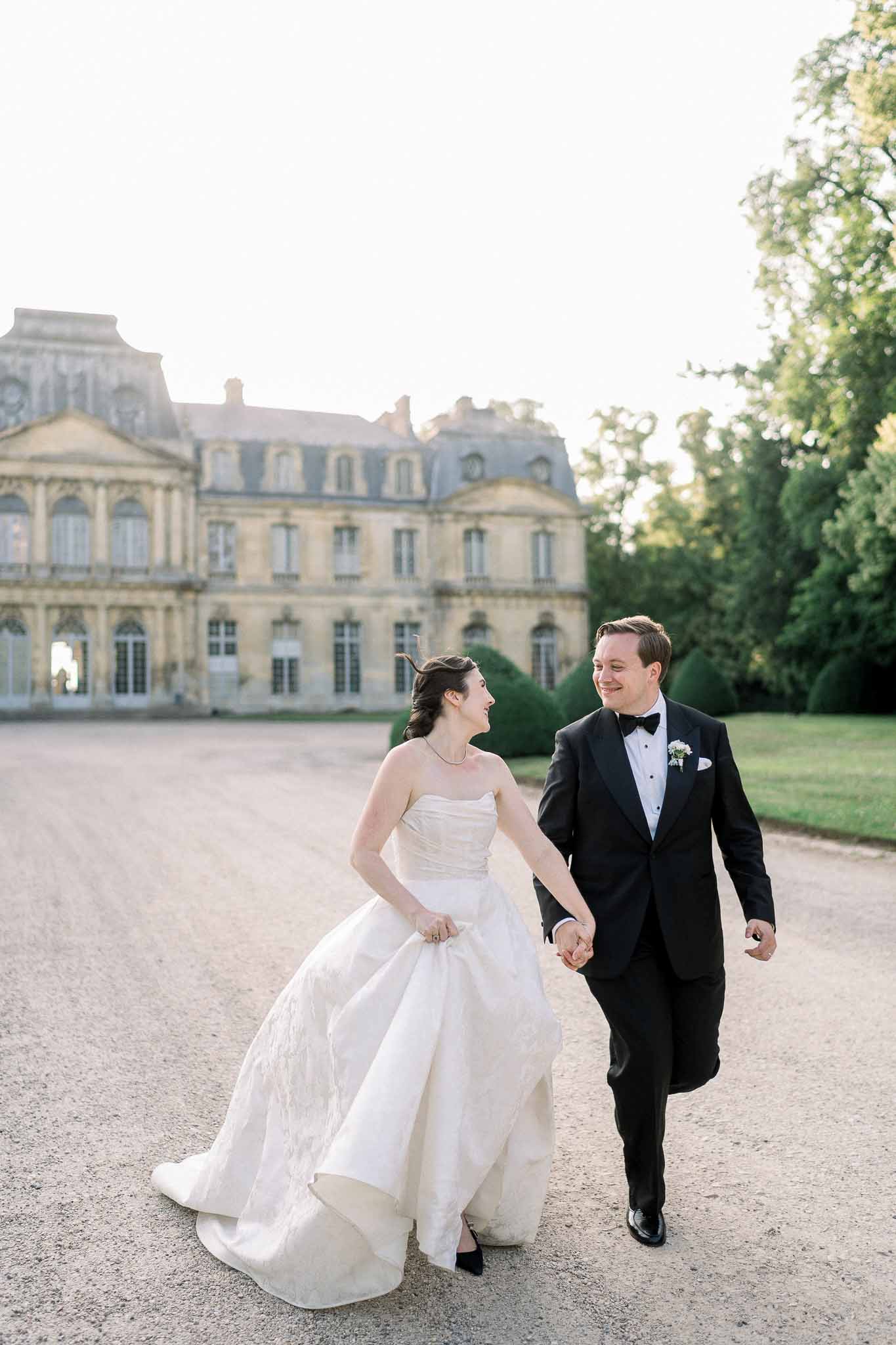 Bride and groom laughing and walking toward camera on chateau forecourt in late afternoon light