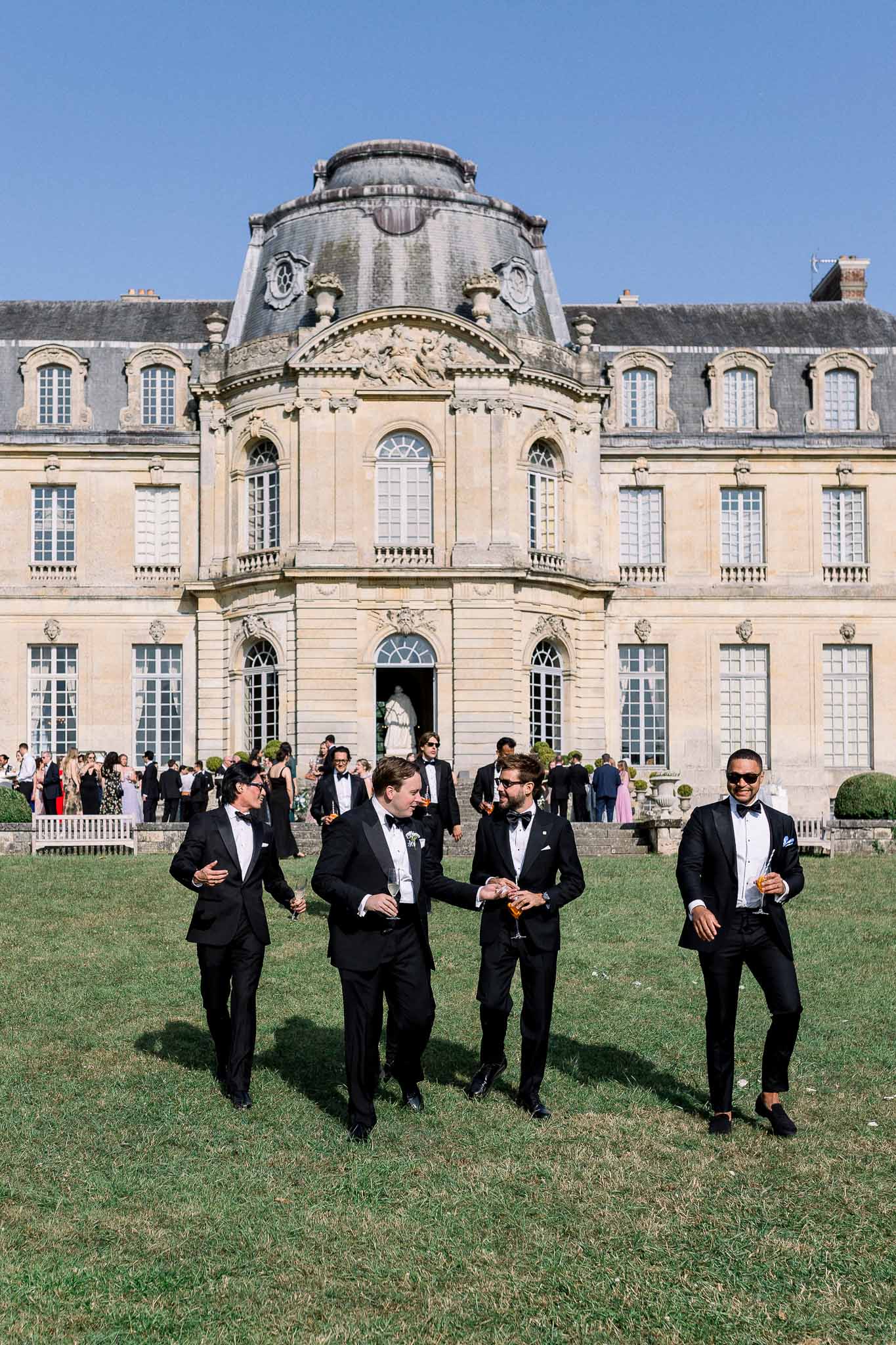 Four groomsmen in tuxedos with champagne walking across lawn before domed classical chateau facade