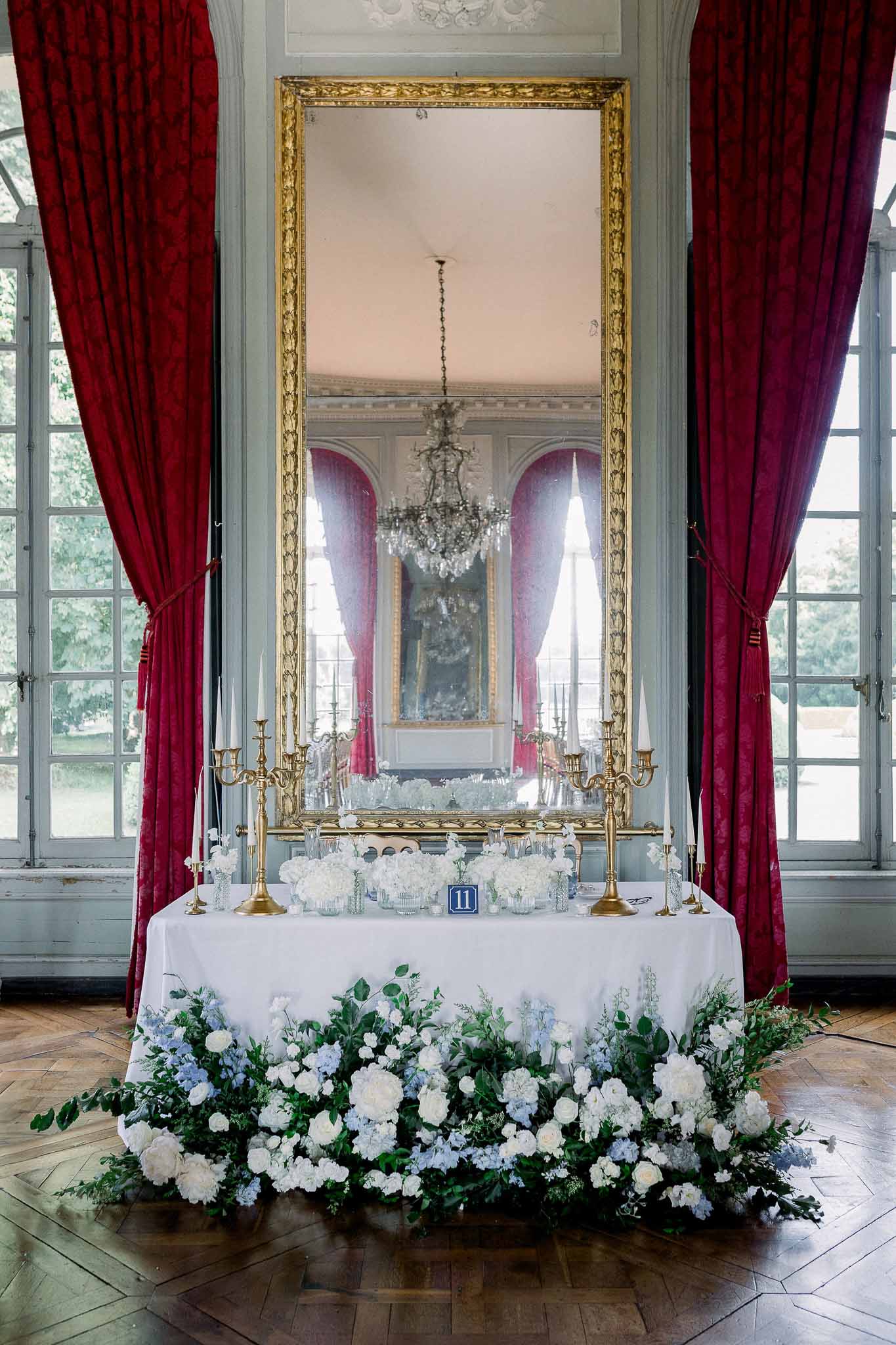 White and blue floral installation beneath gold-framed mirror with crimson curtains in chateau ballroom