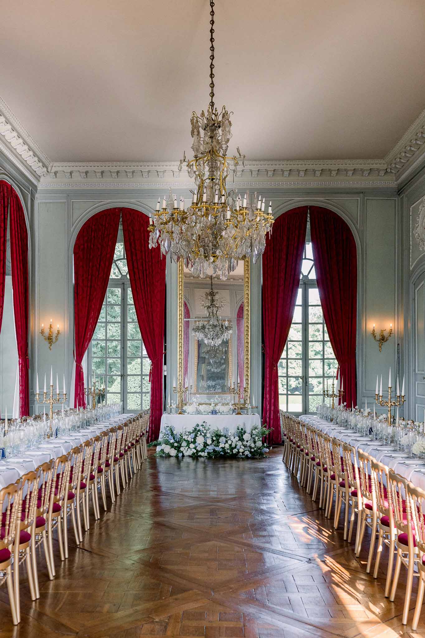 Chateau ballroom with gold Chiavari chairs crimson drapes crystal chandelier and white rose head table
