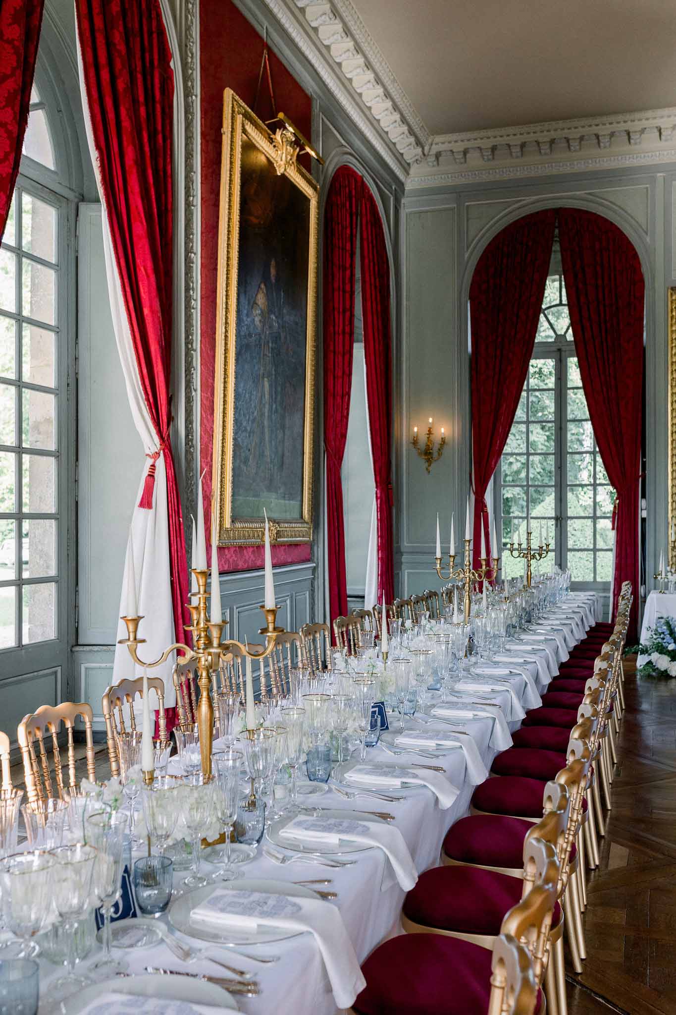 Long banquet table in chateau salon with crimson curtains, gold candelabras, and burgundy velvet chairs