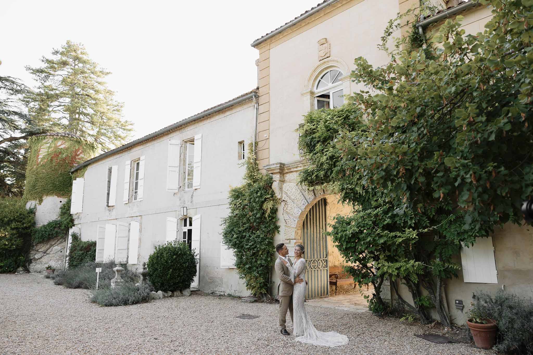 Bride in beaded ivory gown and groom in champagne suit embracing before French bastide entrance