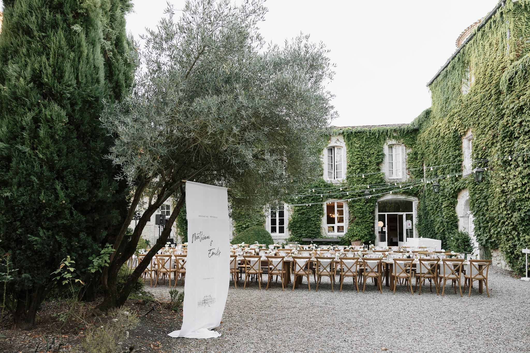 Long tables with white linens and welcome banner before ivy-covered stone manor with string lights on gravel courtyard