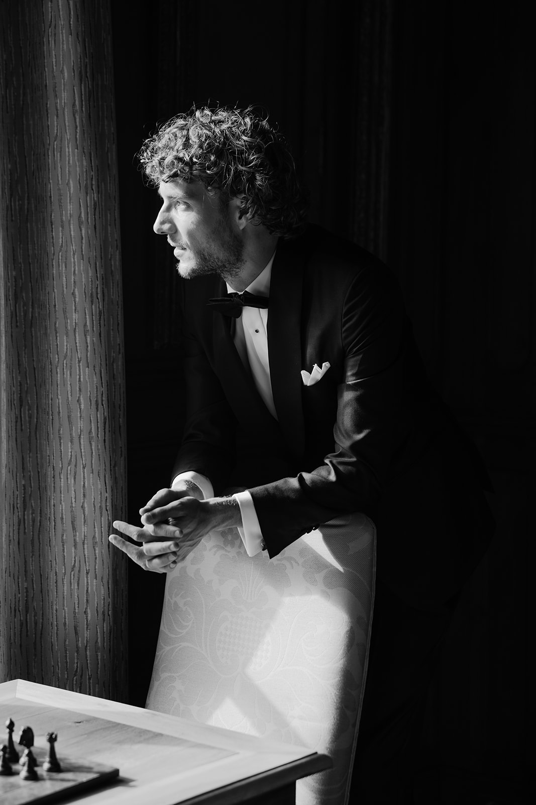 Groom in black tuxedo leaning on damask chair with chess board, dramatic indoor lighting