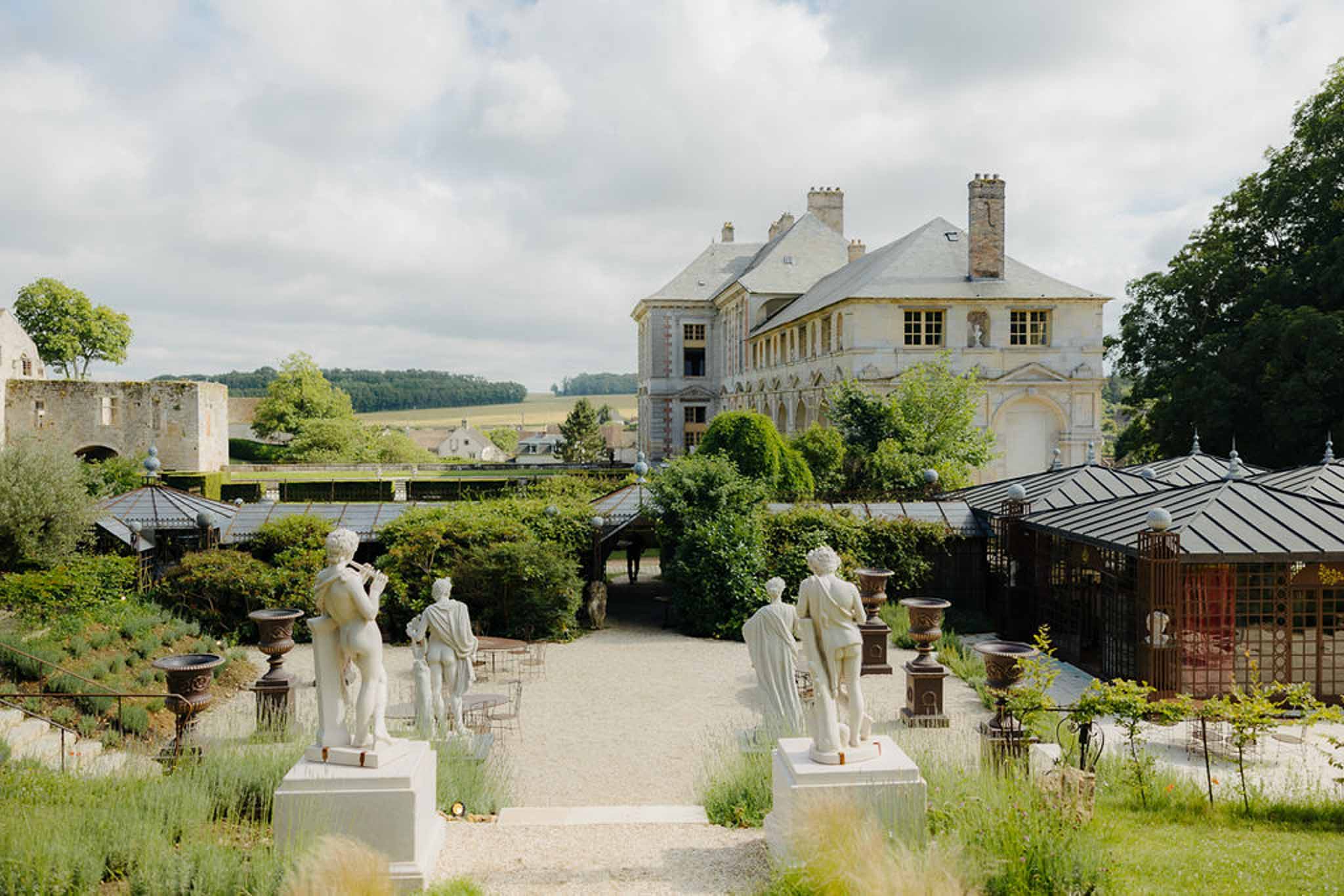 Chateau estate with neoclassical statues lining gravel path, greenhouse wings, and stone ruins at left
