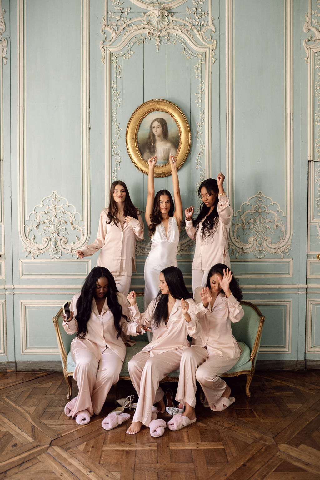 Bride in white satin slip with five bridesmaids in blush pink pyjamas posing on sage settee in blue boiserie room