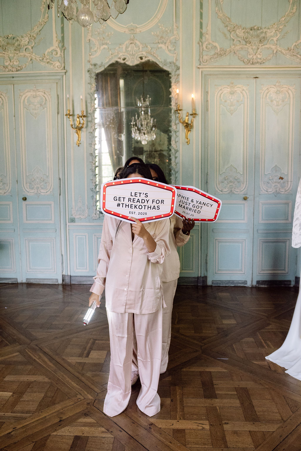 Two women in blush satin pyjamas holding marquee signs in ornate mint-green chateau room