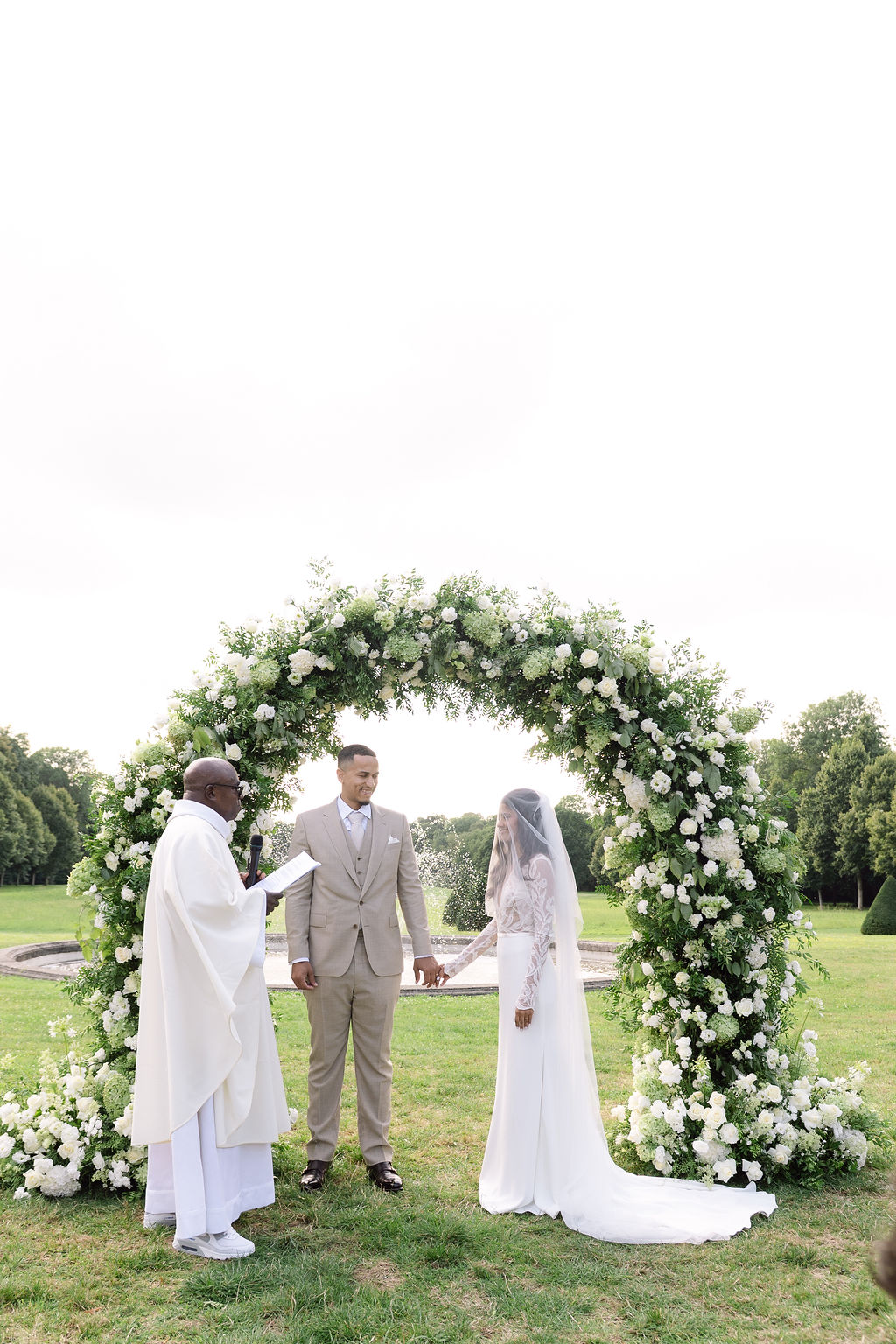 Couple holding hands under circular white hydrangea and rose arch with officiant on lawn before stone fountain