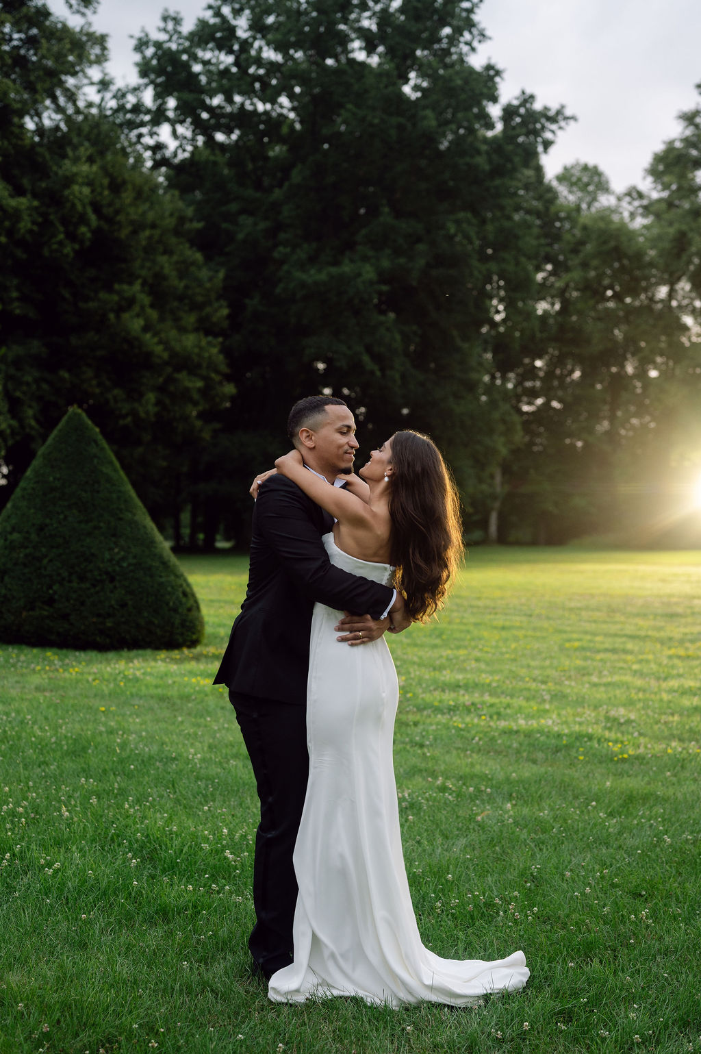 Bride and groom embracing face-to-face on manicured lawn beside conical topiary at golden hour
