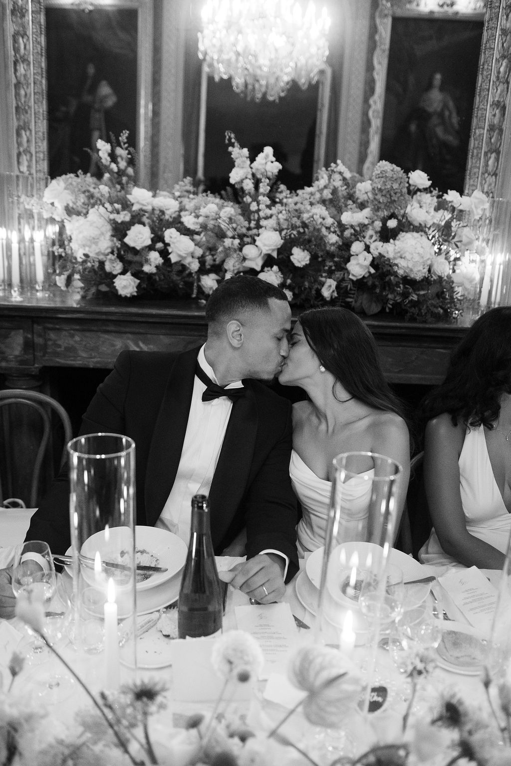 Black and white photo of bride and groom kissing at head table during chateau reception with floral mantelpiece