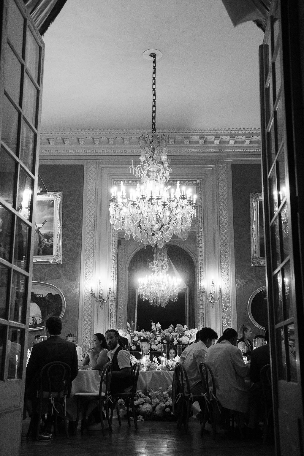 Black and white of candlelit ballroom reception through tall doors with crystal chandeliers and cascading floral runner