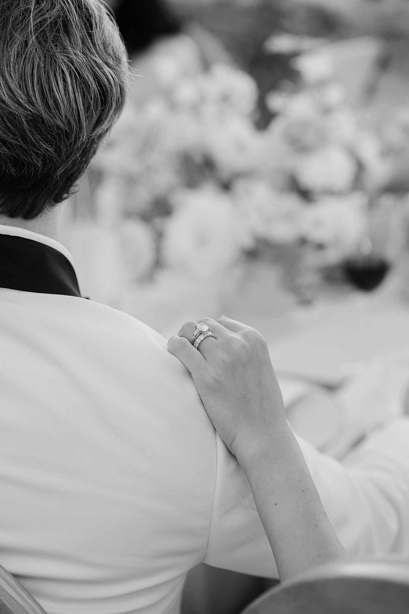 Black and white close-up of bride's hand with oval solitaire and diamond band resting on groom's shoulder