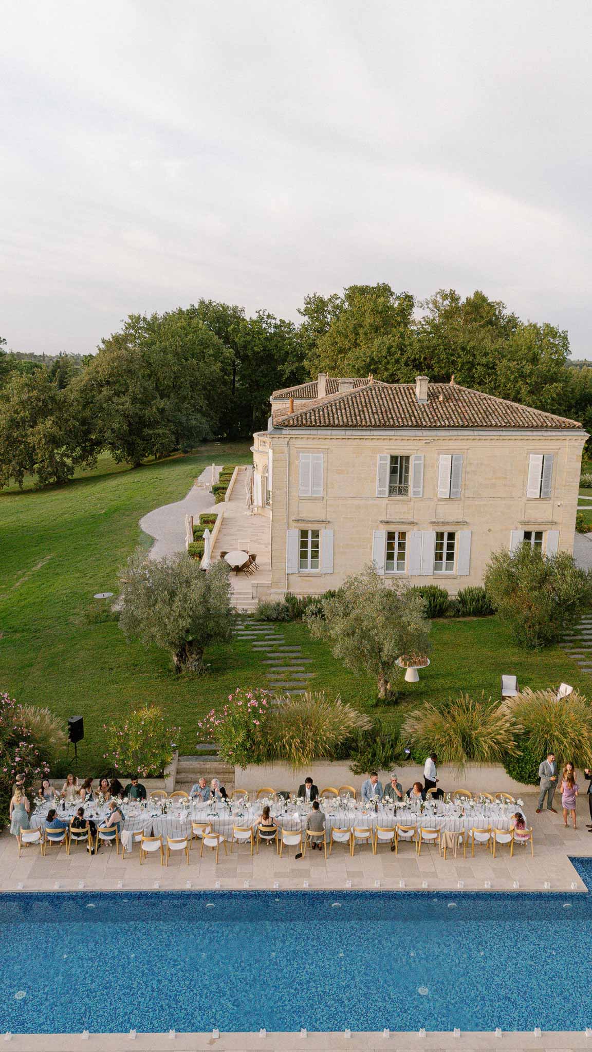 Aerial reception dinner along pool with white floral tables and gold chairs before limestone manor with terracotta roof