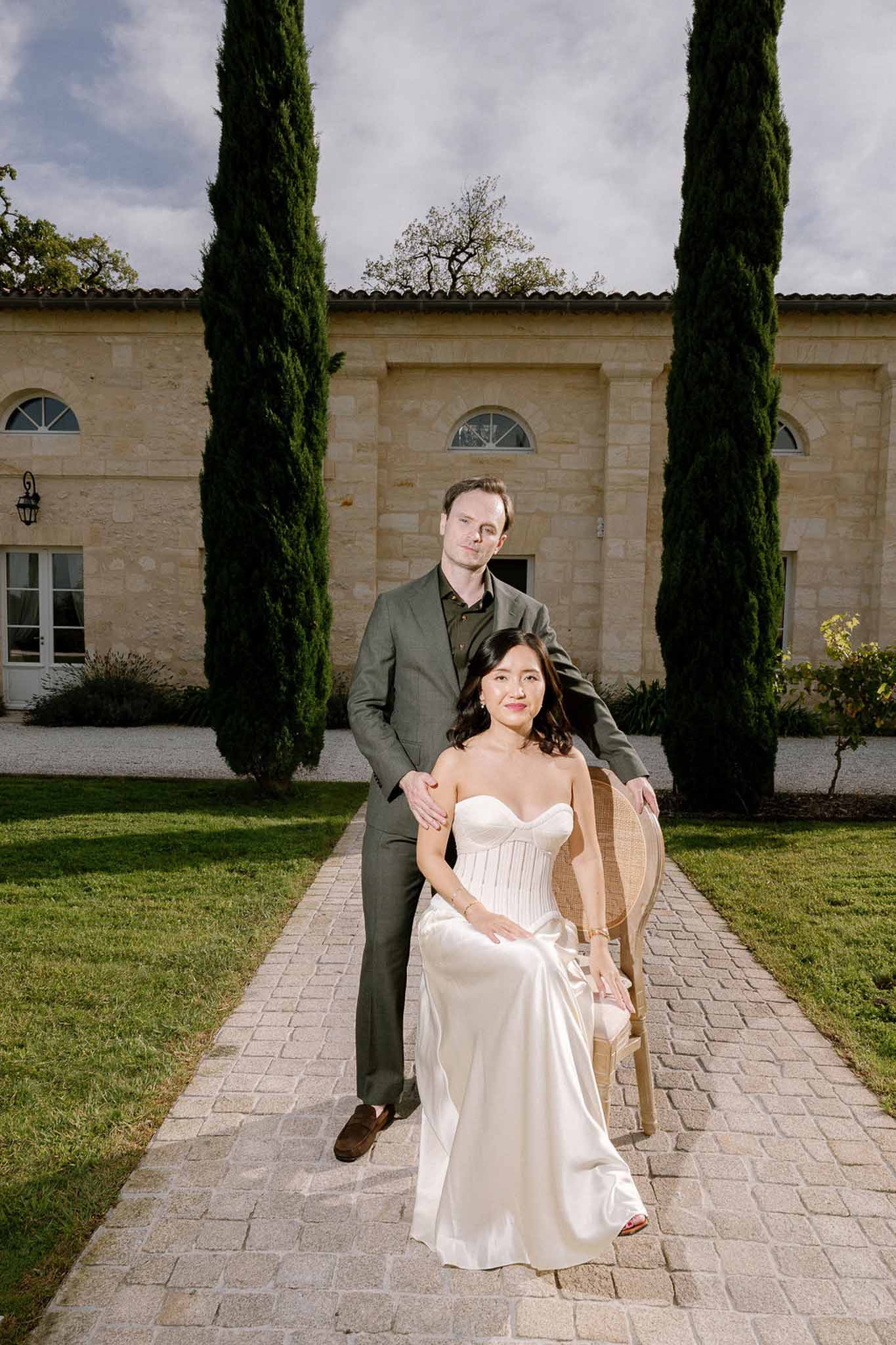 Bride seated in cane chair with groom standing behind her before limestone chateau with cypress trees