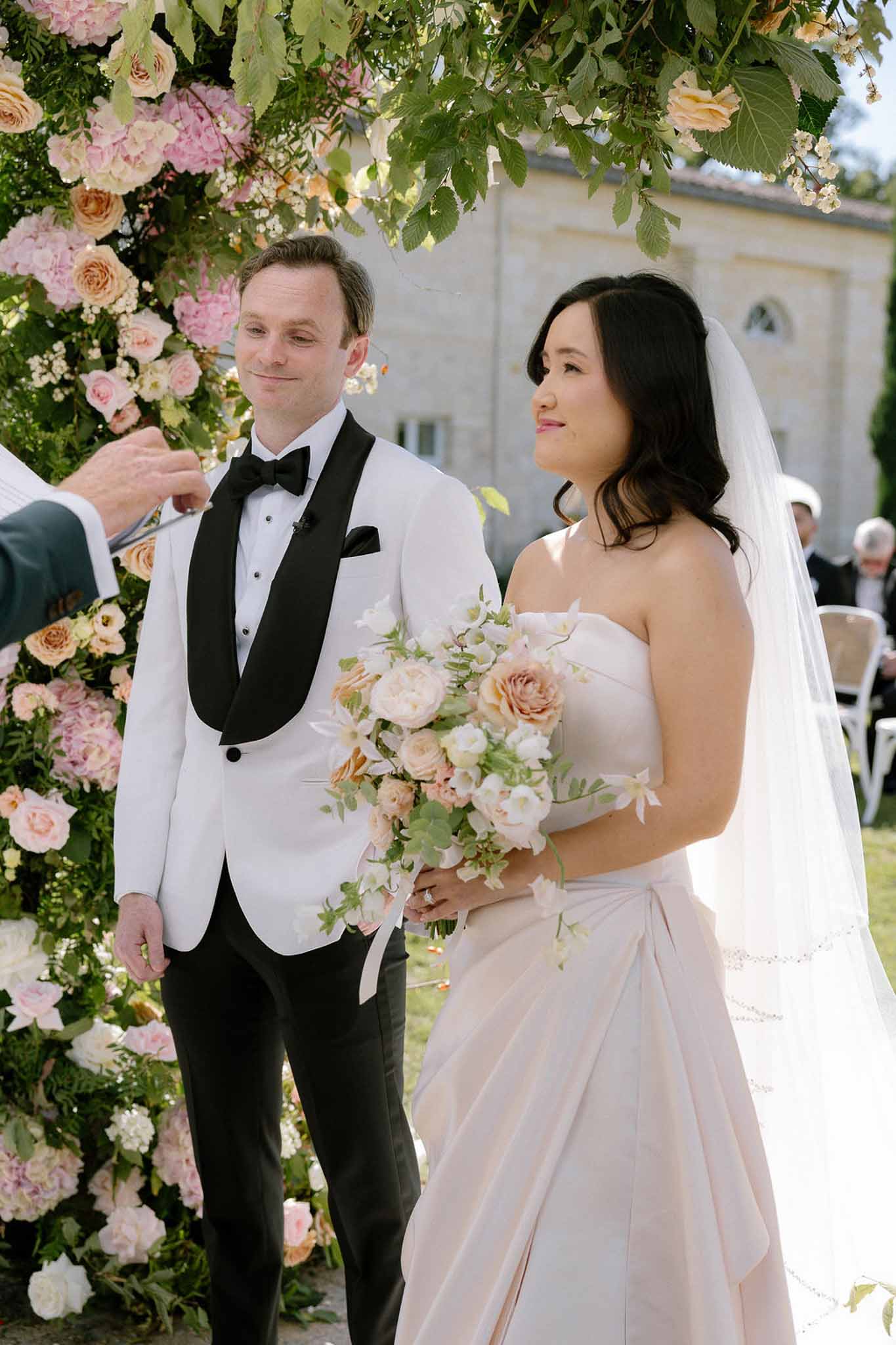 Bride in blush gown and groom at altar beneath dense blush and peach hydrangea floral arch