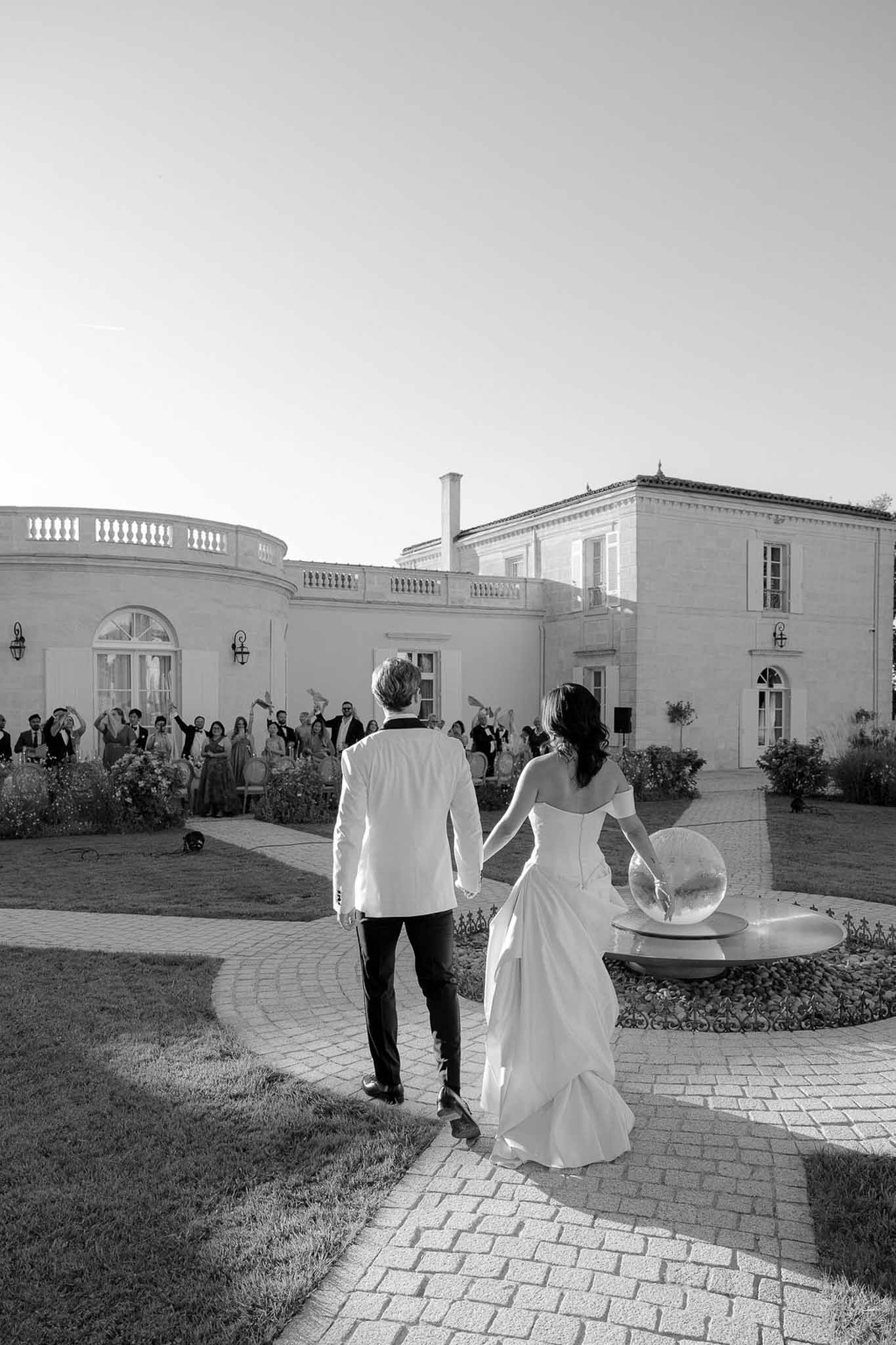Bride and groom walk toward chateau terrace as 30 guests cheer from balustrade wing in B&W