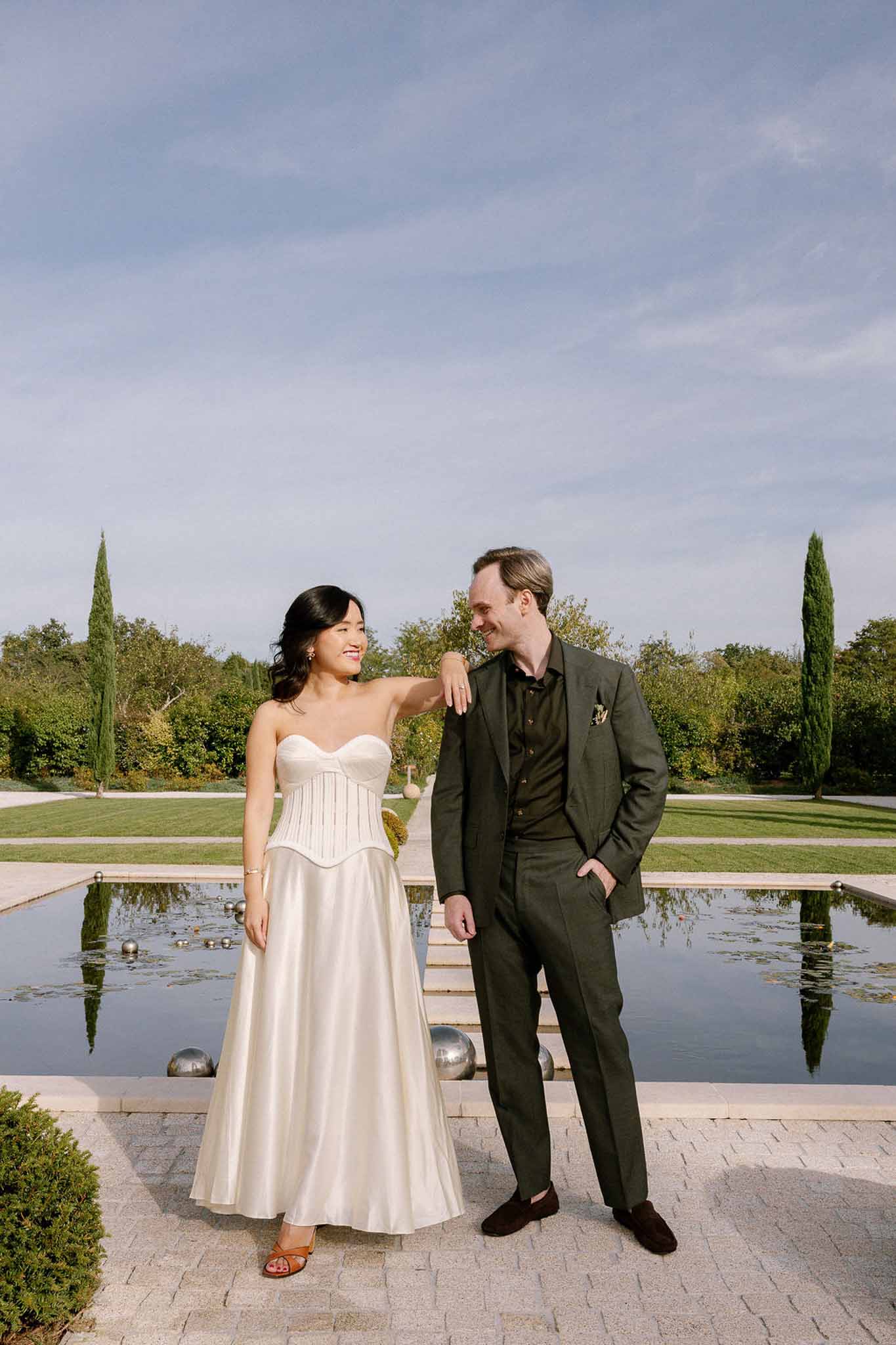 Bride in strapless ivory gown and groom in green suit posing by reflecting pool at French chateau garden