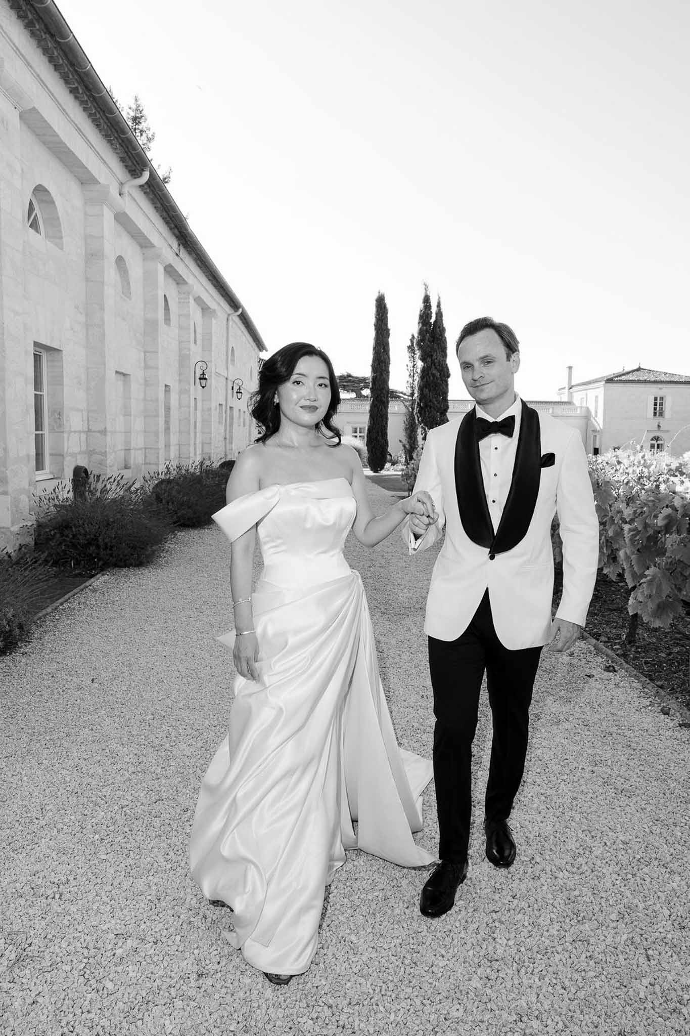 Black-and-white portrait of bride and groom holding hands walking on gravel path at French estate