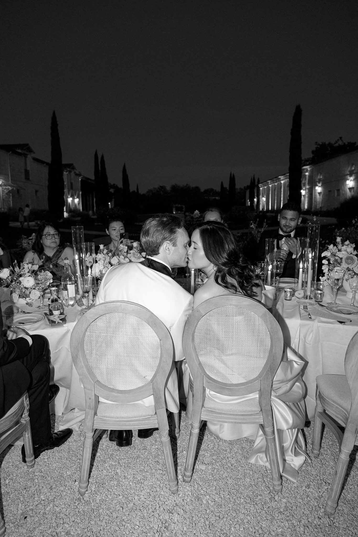 Black and white evening reception with couple kissing at candlelit round table before cypress trees