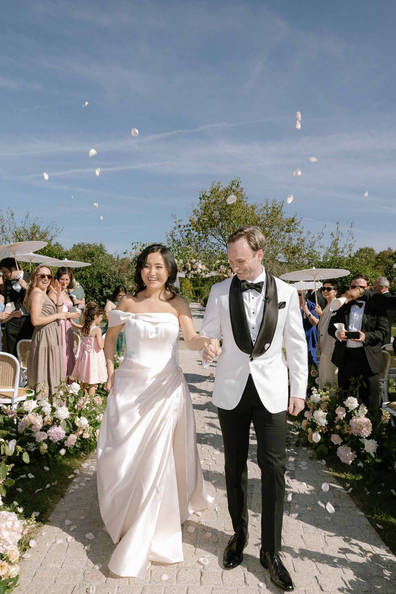 Couple walks through rose petal toss past blush peony aisle arrangements with guests holding parasols