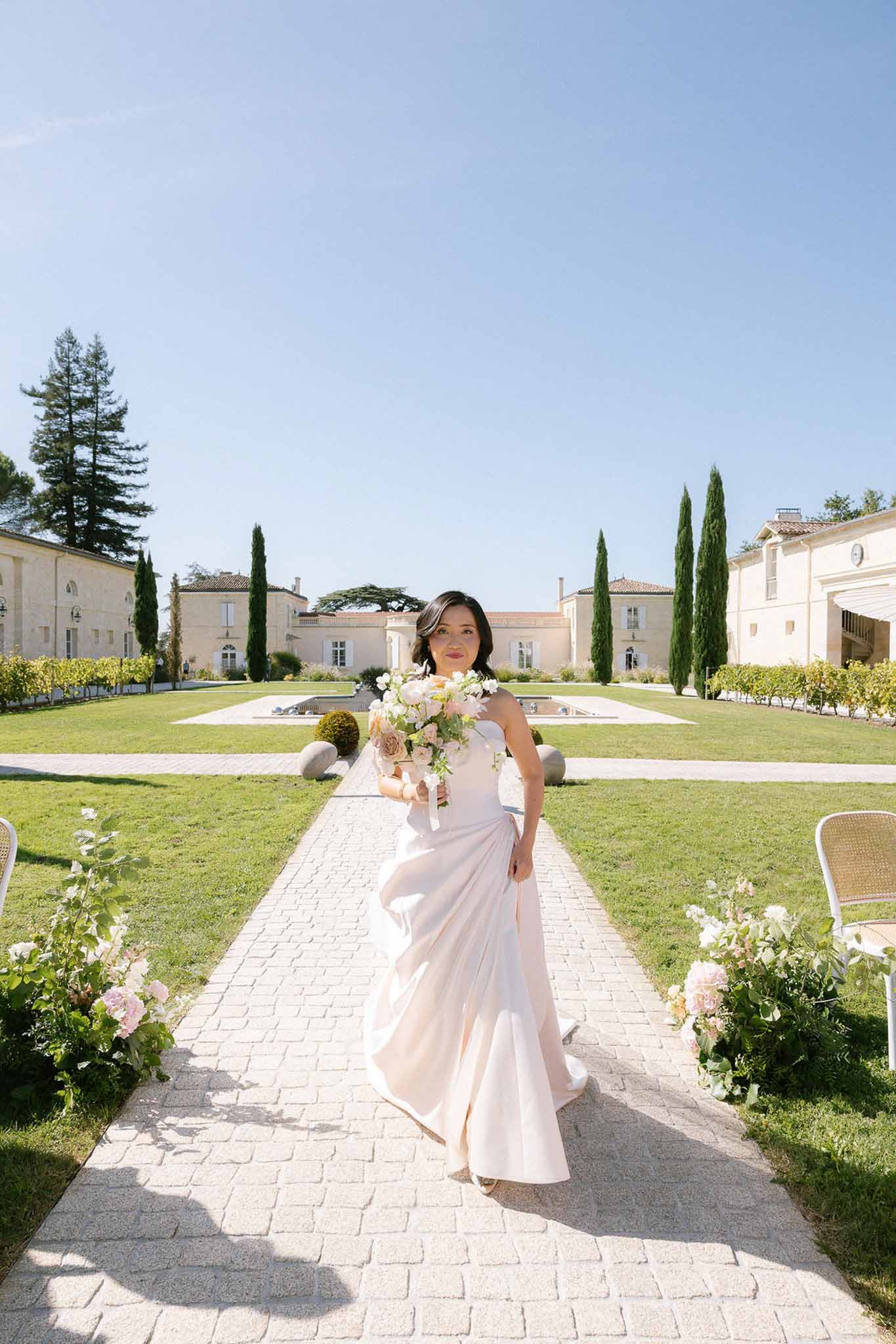 Bride in blush strapless gown walking down outdoor aisle with rose arrangements toward French chateau