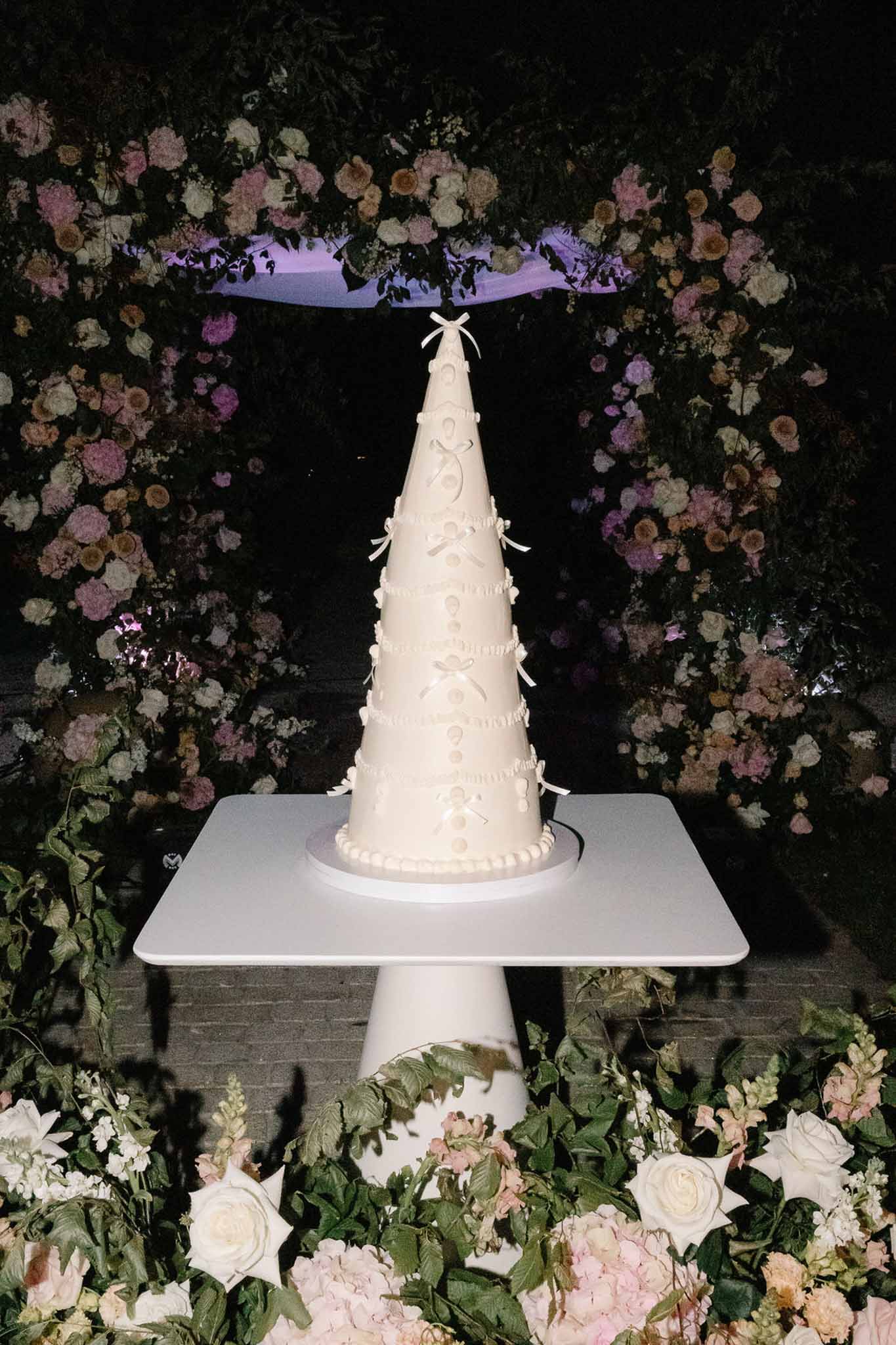 Cone-shaped ivory wedding cake with pearl dots and ribbon bows displayed against a blush and mauve floral arch at night